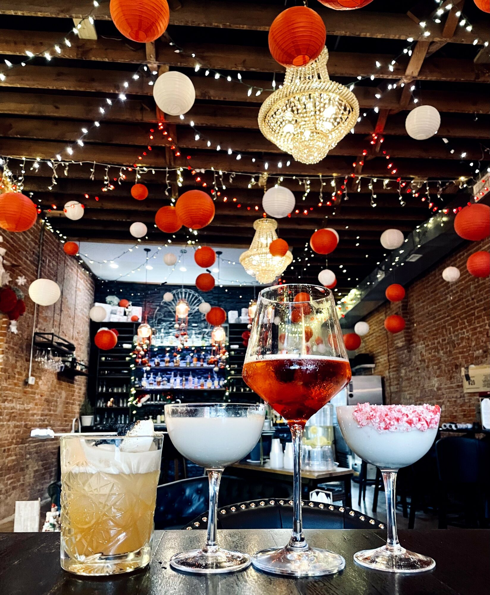 Decorative ceiling with hanging lanterns and string lights, bar with drinks on table, brick walls, cozy ambiance.