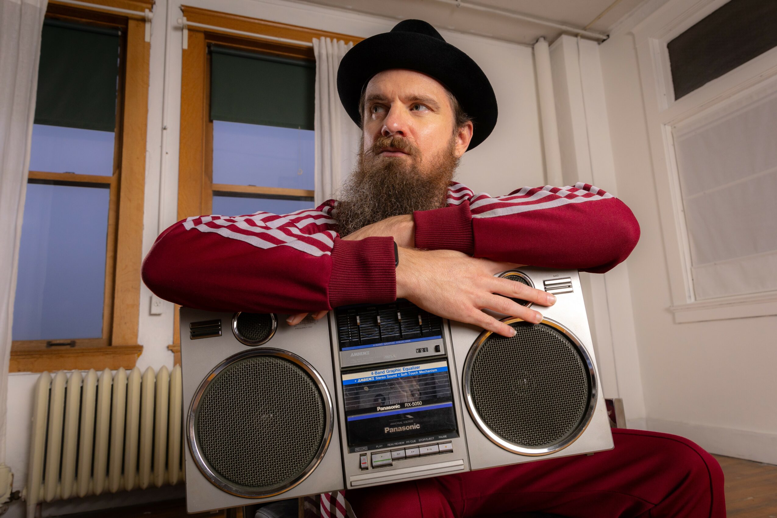 Man with a beard and hat leaning on a large boombox in a room with windows and white walls.