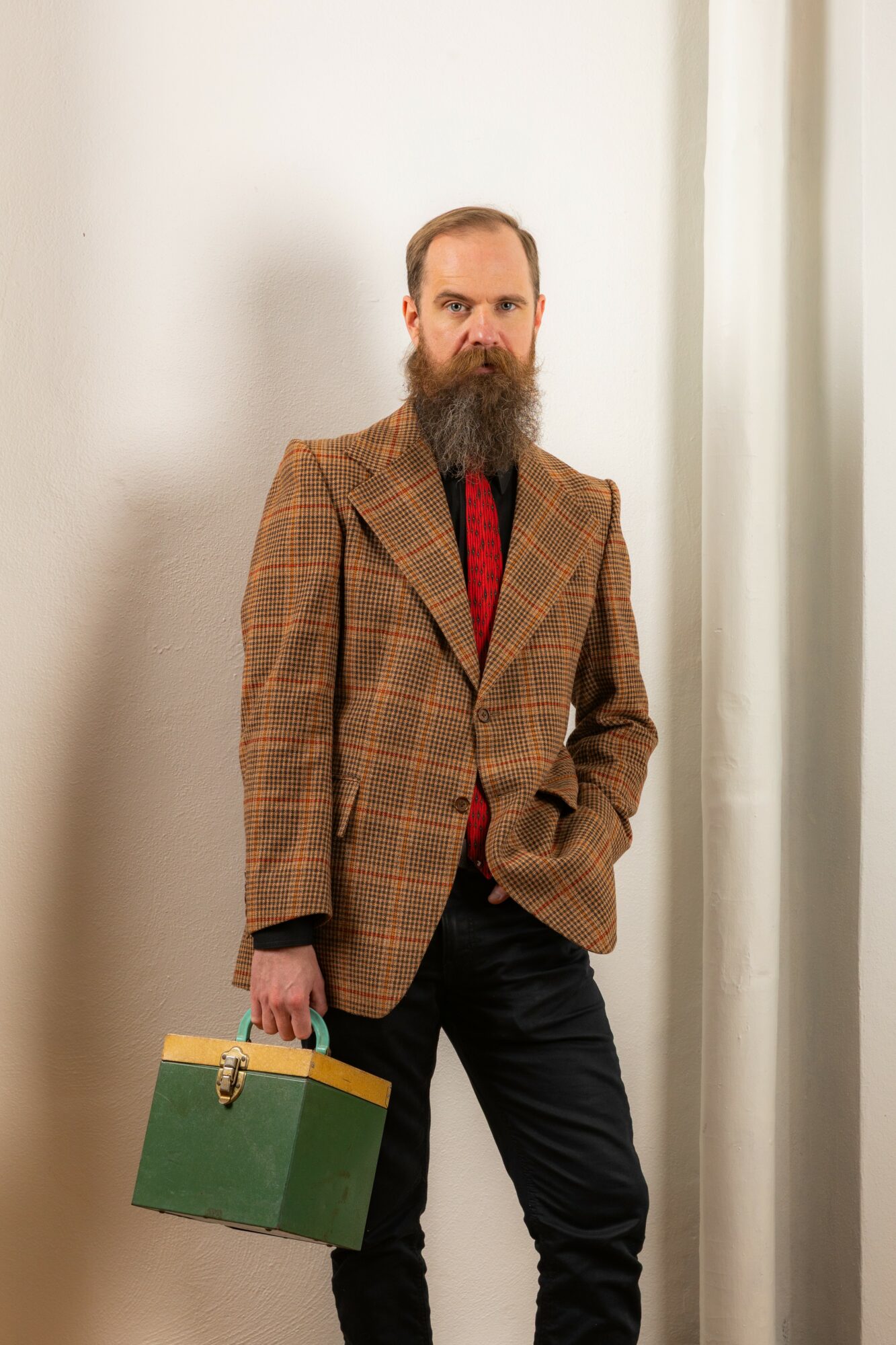 Man with a beard in a plaid blazer holding a green and yellow briefcase, standing indoors near a white wall and curtain.