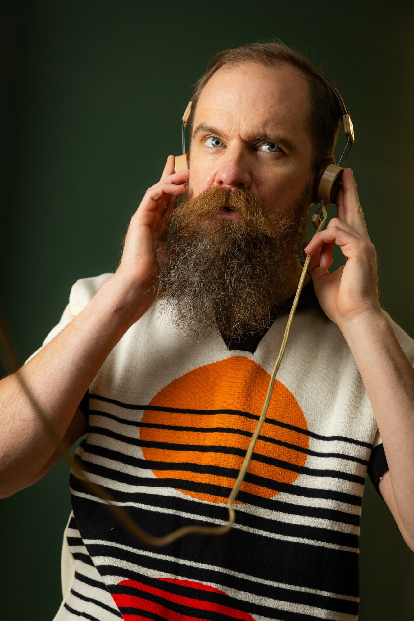Man with a beard and mustache wearing headphones, looking surprised, against a dark green background.