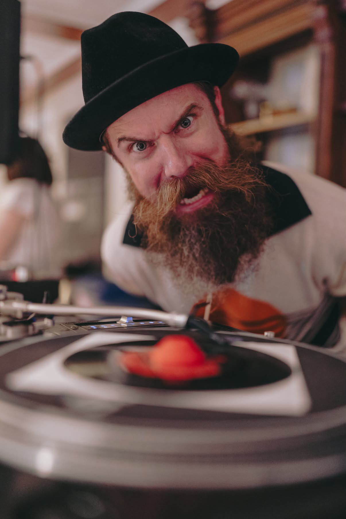 Man with a beard and hat looks angry at a record player with a red object on the vinyl.