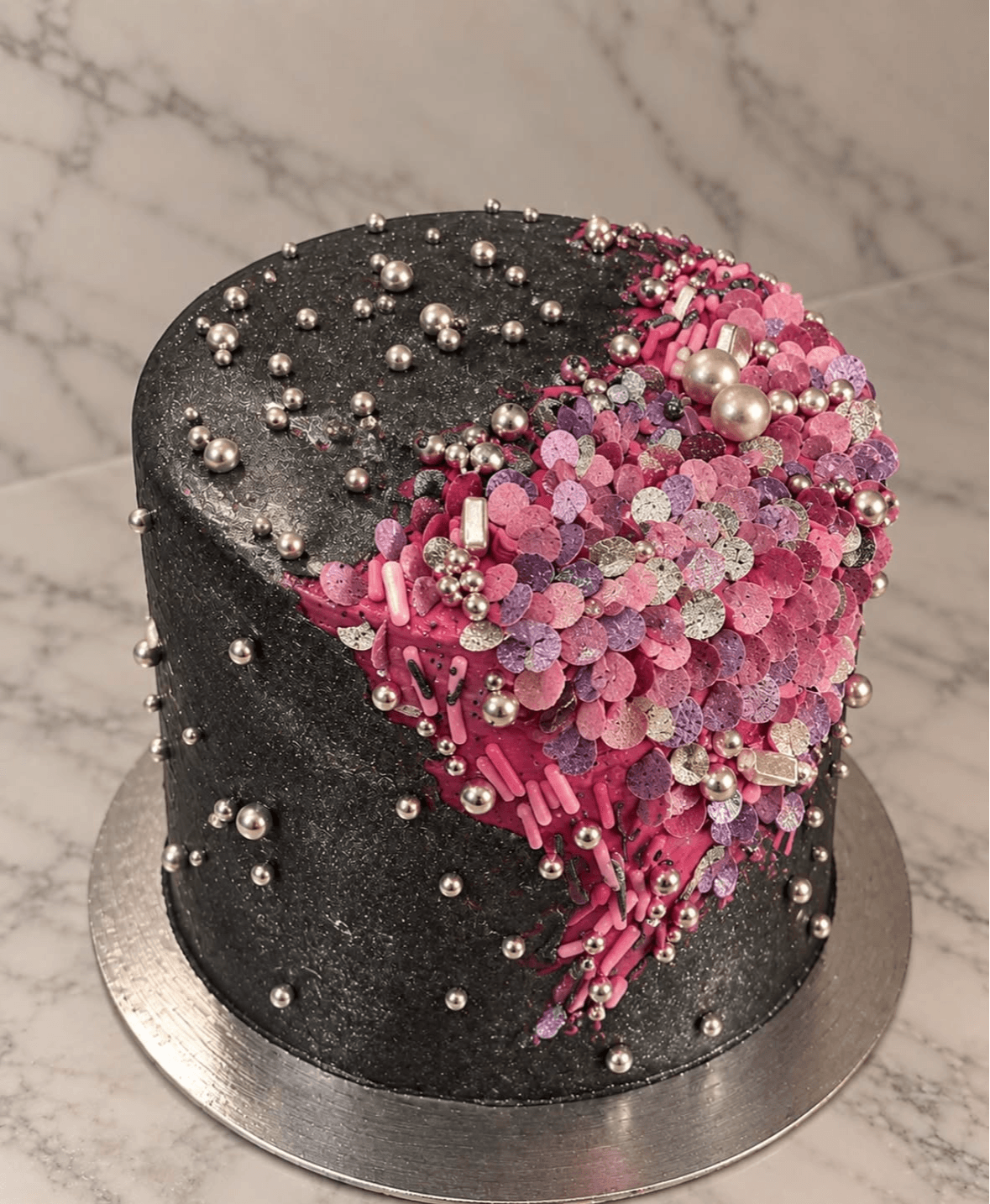 A black cake with silver and pink decorations, including pearls and glitter, on a silver base against a marble background.