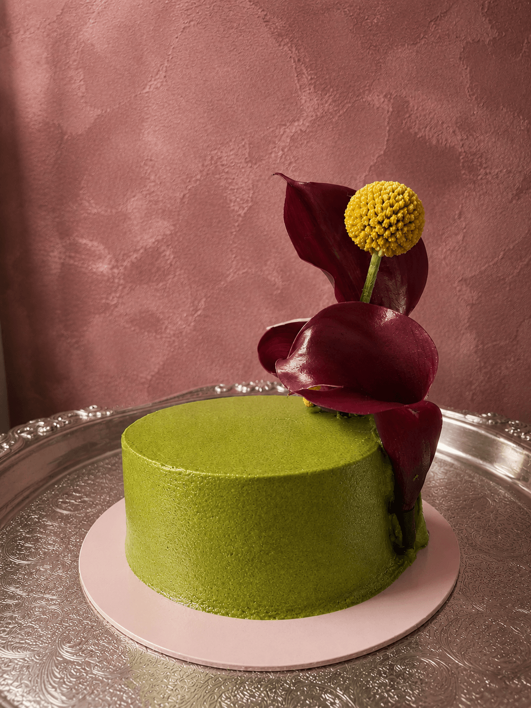 Round green cake with dark red flower decoration on top, placed on a silver tray against a pink wall.