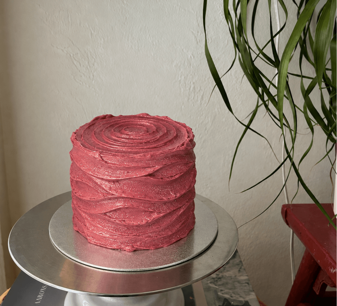 Pink layered cake on a silver stand, with a plant and a red chair nearby, against a plain wall.