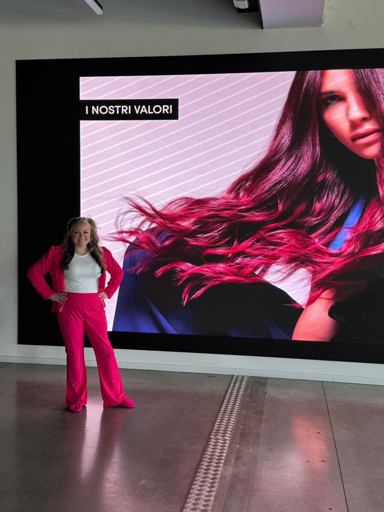 Person standing in front of a large digital screen displaying a woman with long, wavy hair wearing a black outfit and pink pants.