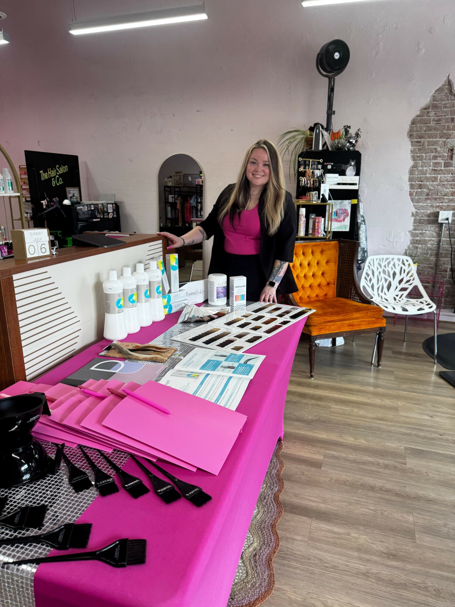 Woman standing behind a table with skincare products, pink folders, and promotional materials in a room with wooden flooring.