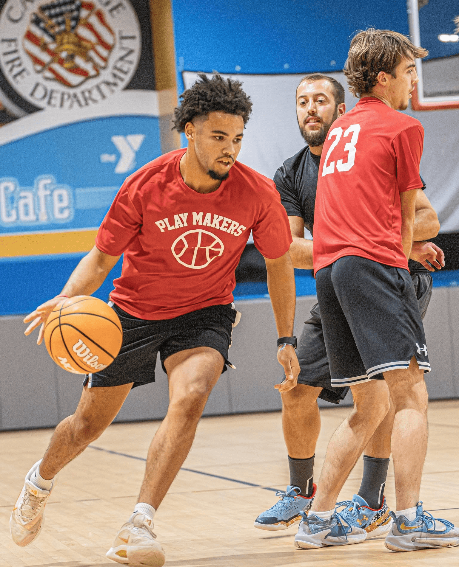 Three young men playing basketball on an indoor court, one dribbling the ball, others standing nearby, with a logo in the background.