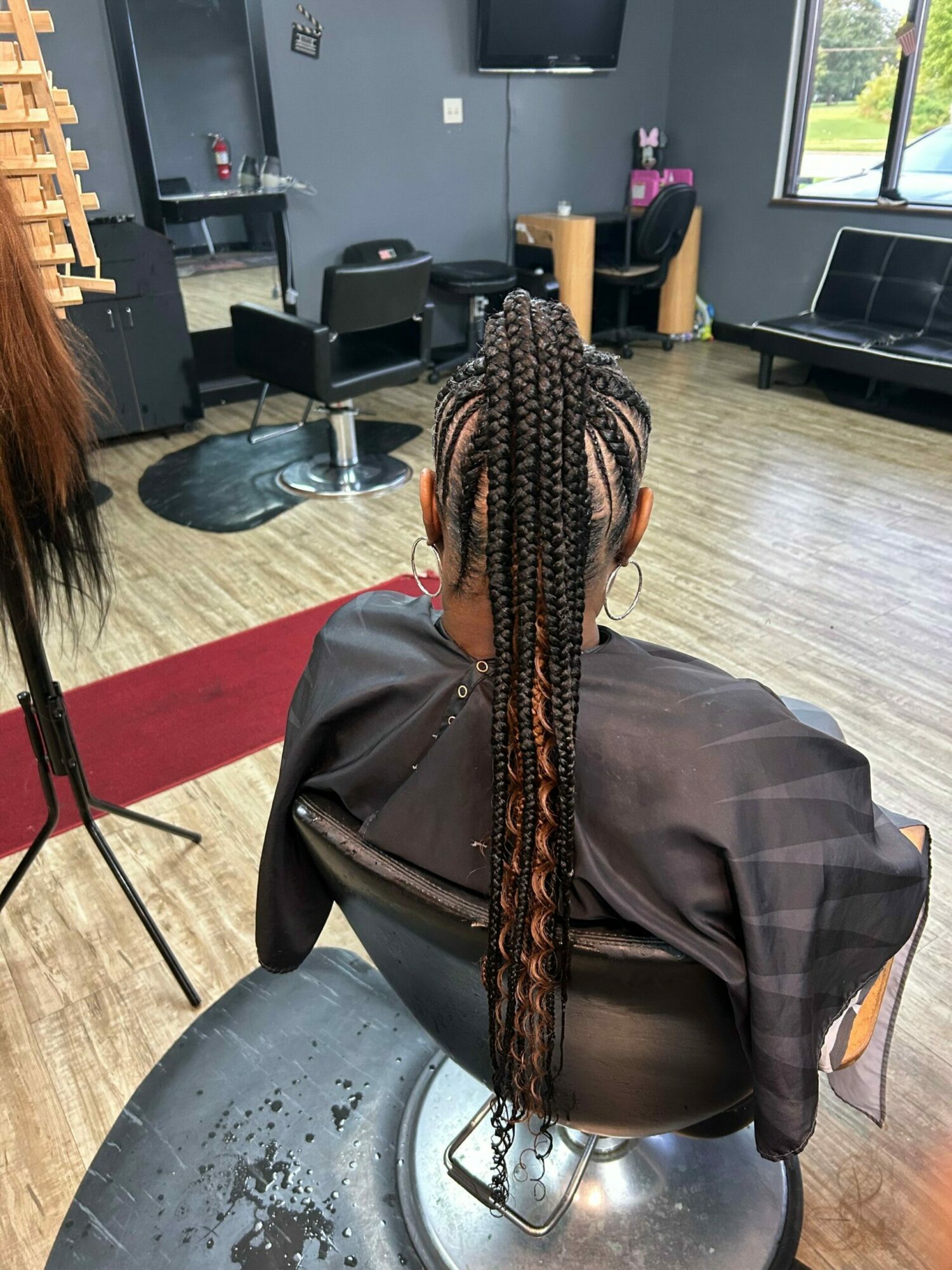 Person with long braided hair sitting in a salon chair, facing away, with hairdresser working nearby in a salon setting.