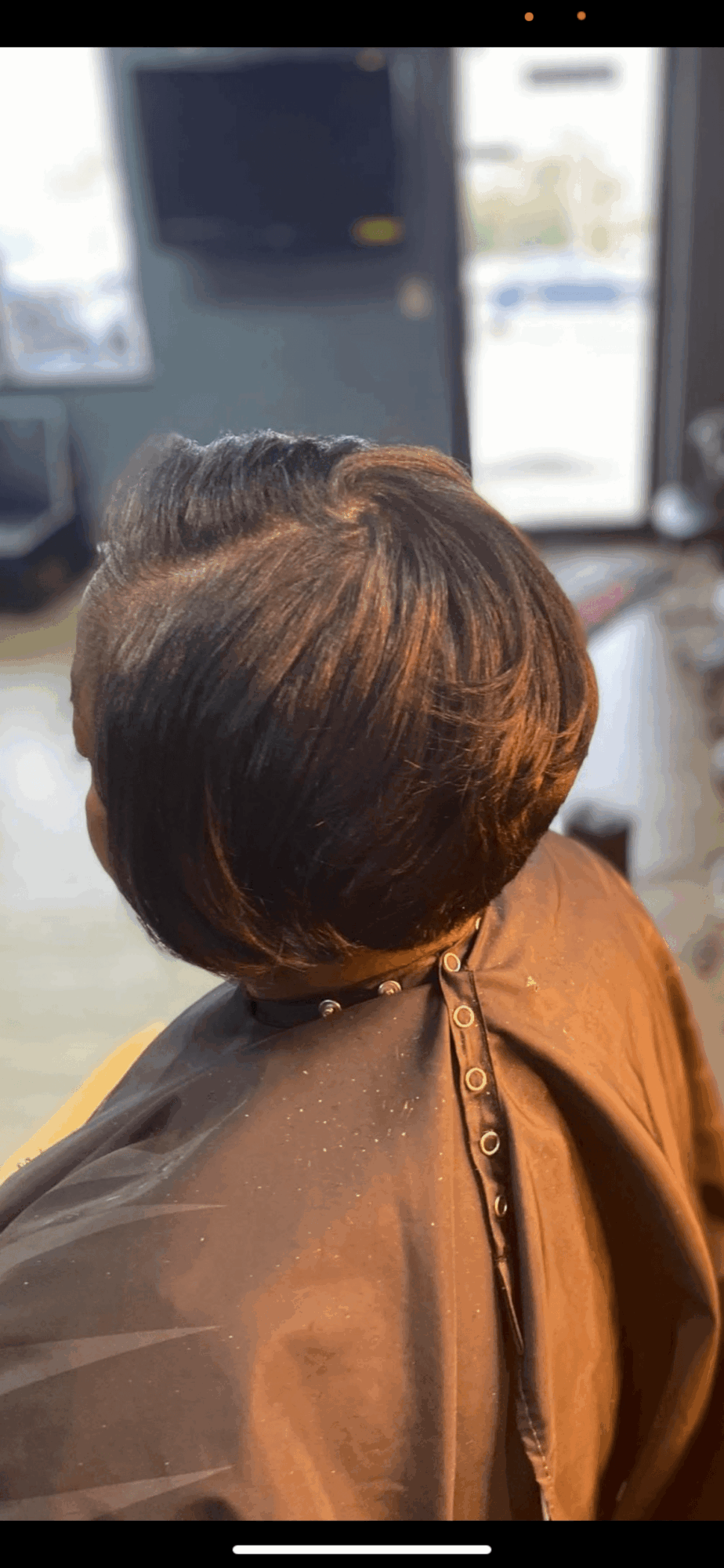 Back view of a person with short, layered brown hair in a salon chair, wearing a brown cape, with a window and TV in background.