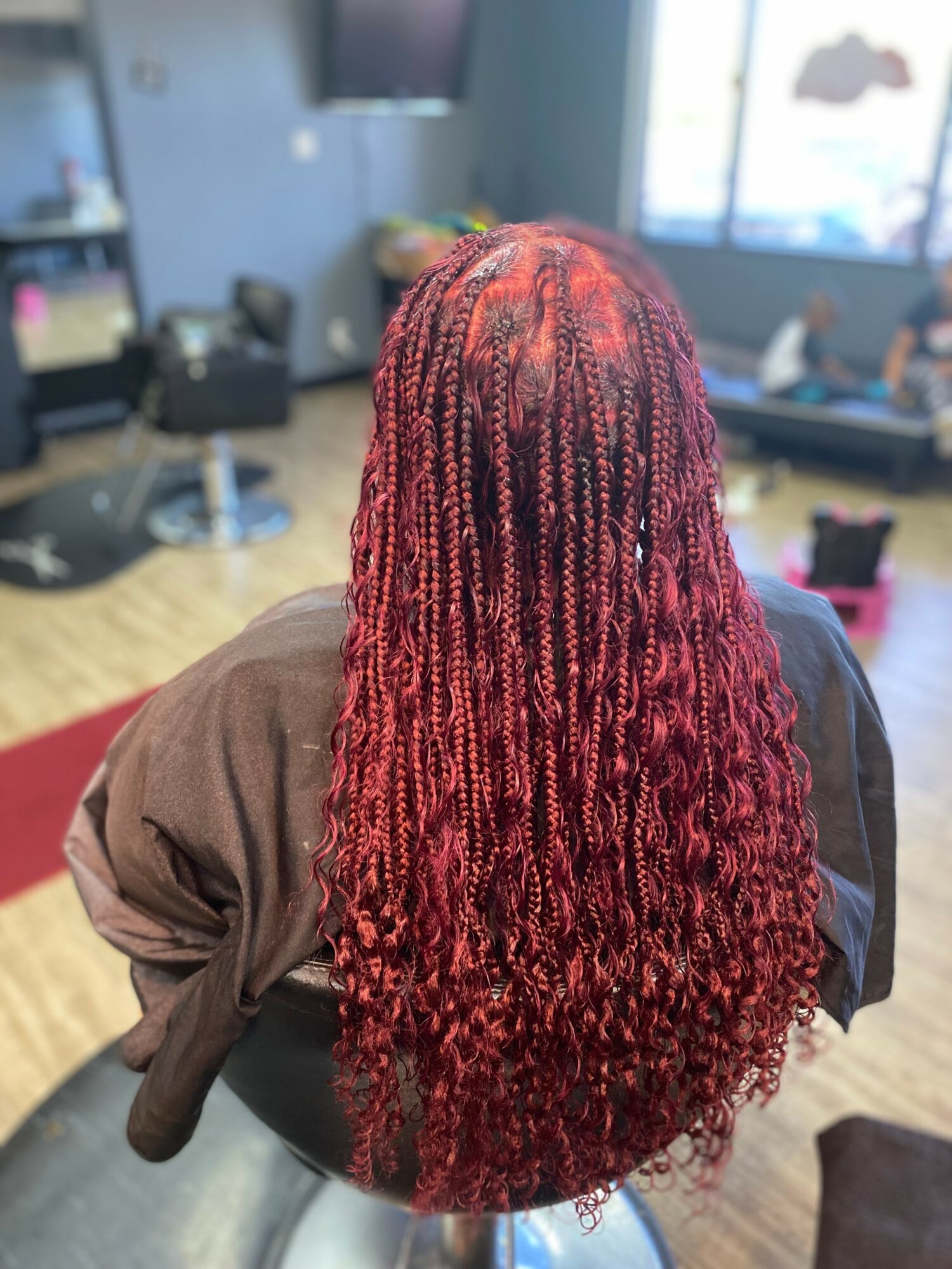 Person with long, red, curly braids sitting in a salon, viewed from behind, with salon chairs and windows in the background.