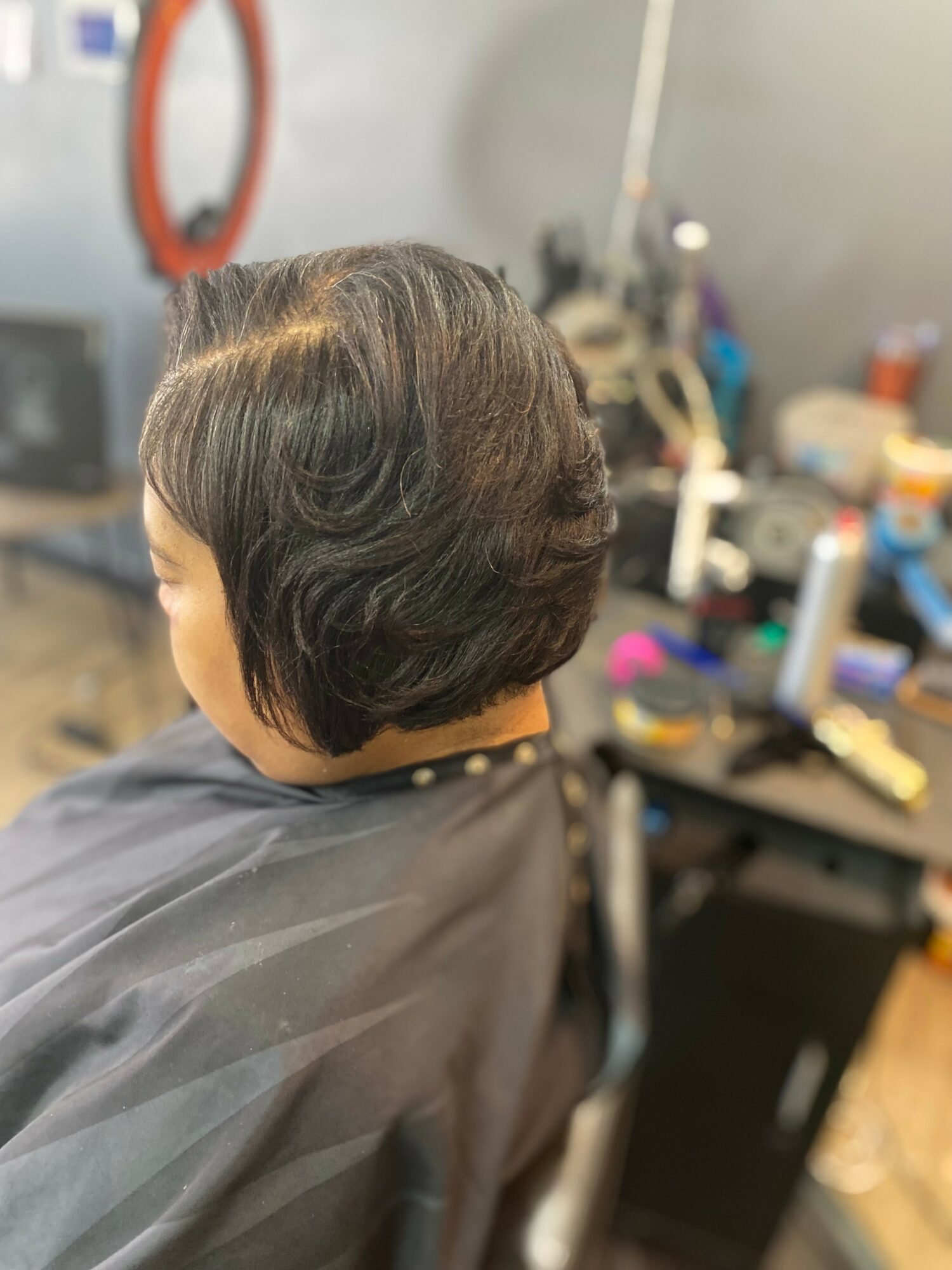 Back view of a person with short, dark, wavy hair in a salon chair, with hairdressing tools and products in the background.