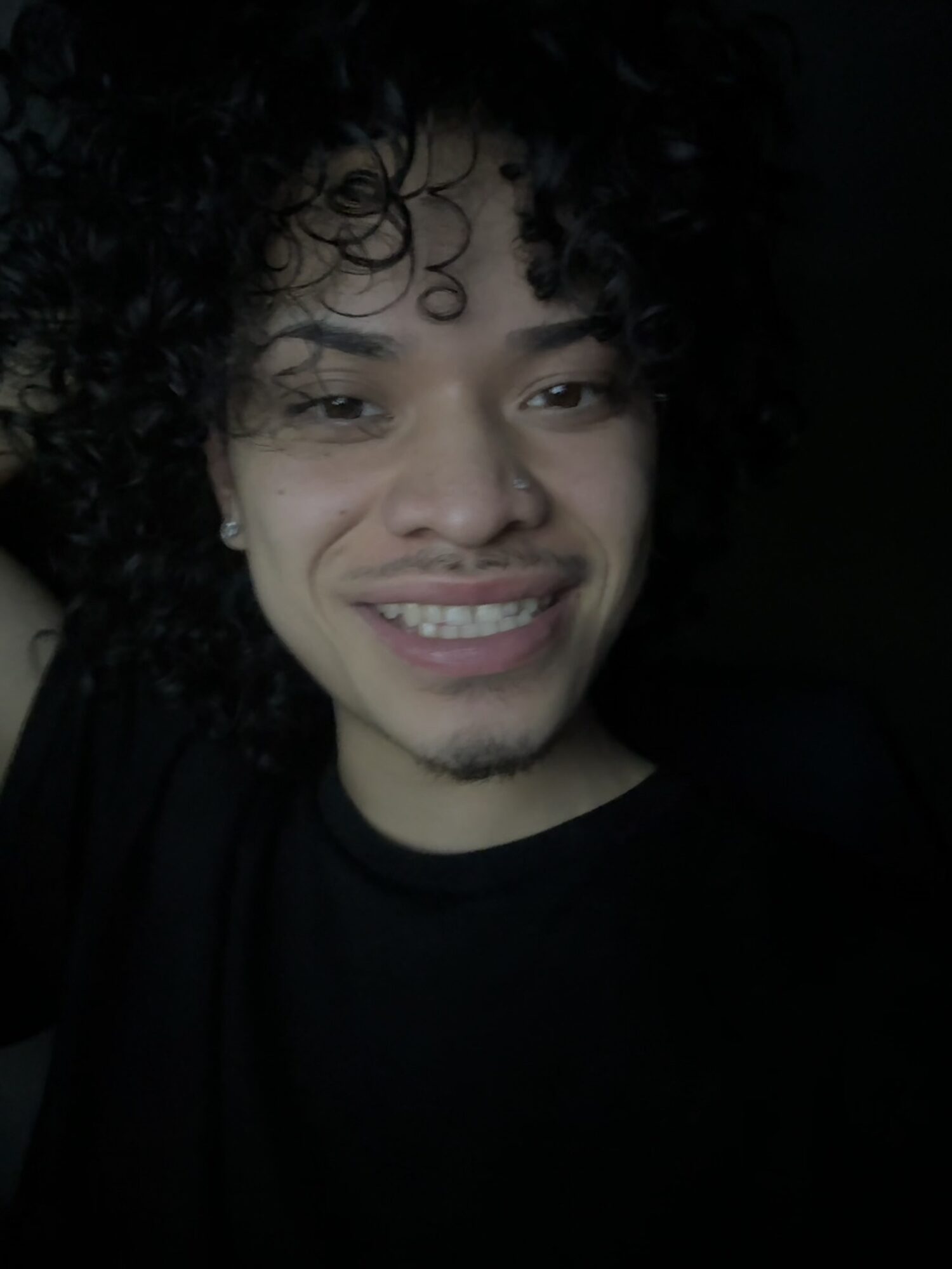 Young person with curly hair smiling, wearing a black shirt, dark background.