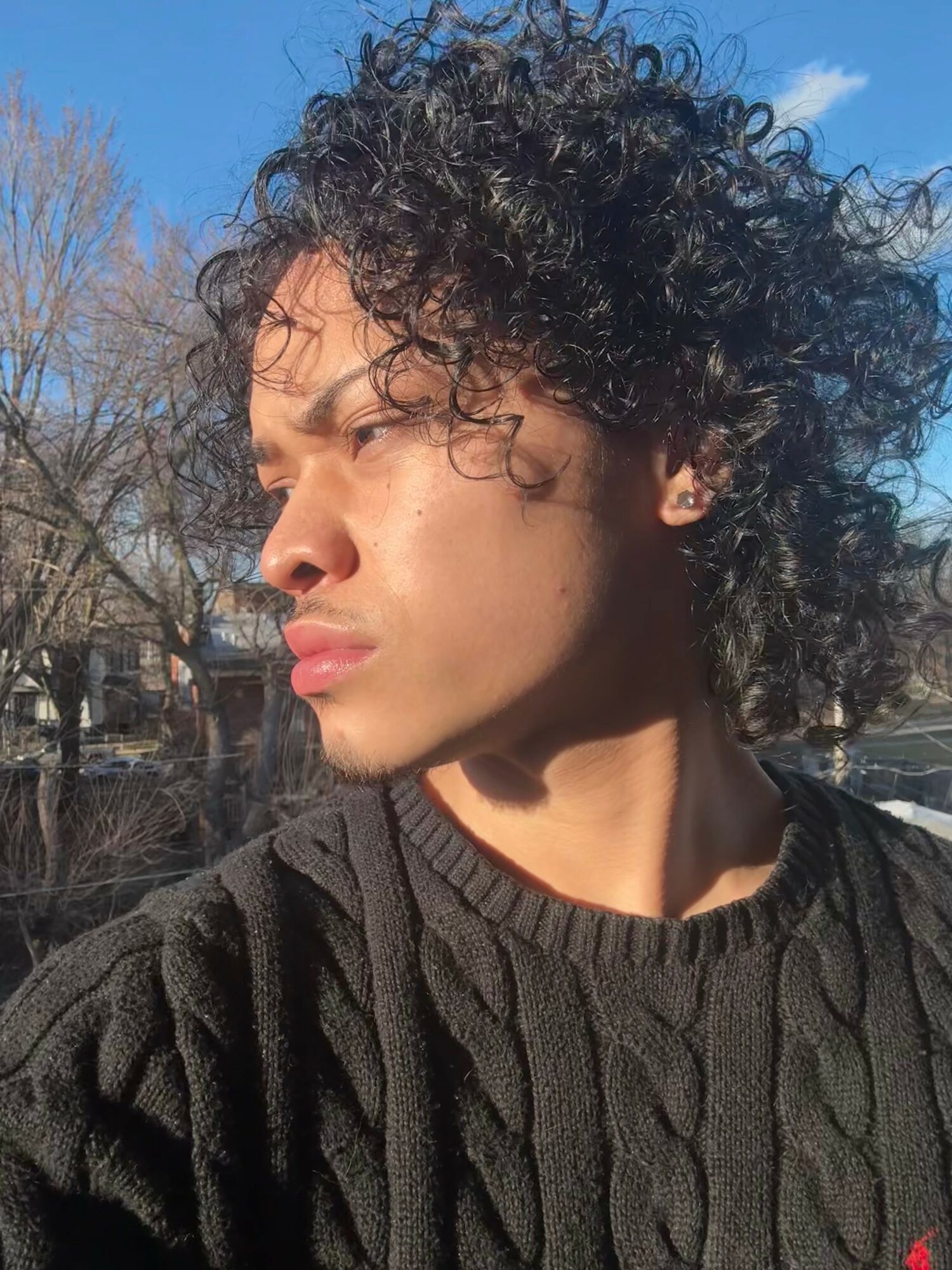 Young person with curly hair looking to the side outdoors on sunny day, wearing a black sweater.