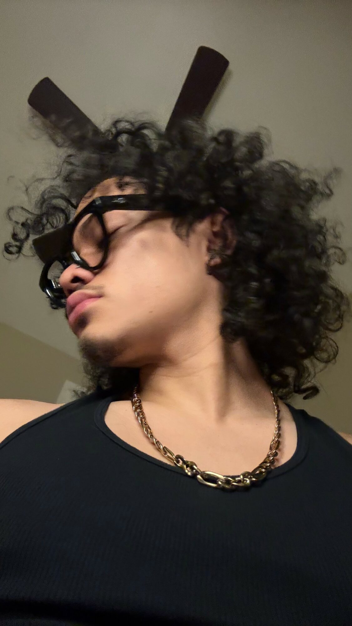 Person with curly hair, glasses, and a chain necklace, looking to the side, ceiling fan above.