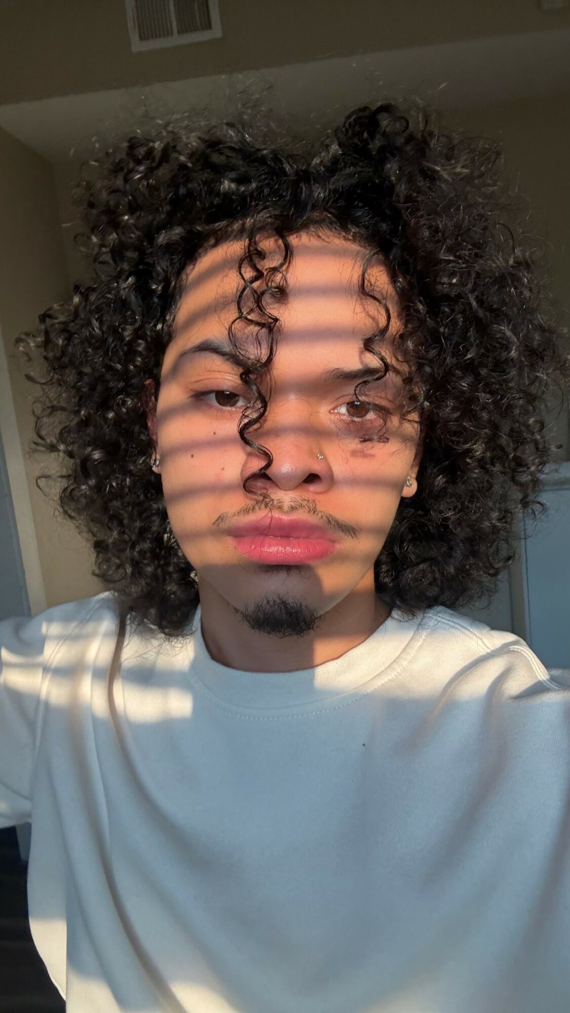Person with curly hair and light skin, sunlight casting shadows through blinds, wearing a white shirt, indoors.