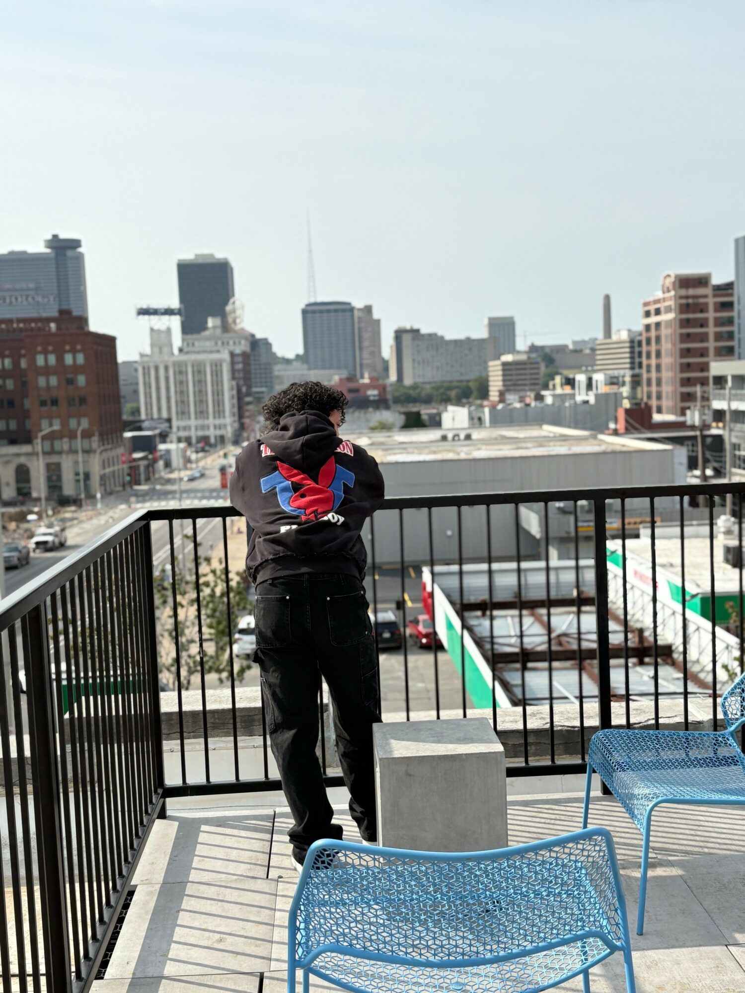 Child with curly hair stands on a rooftop balcony overlooking a city skyline, facing away from the camera, with arms crossed.