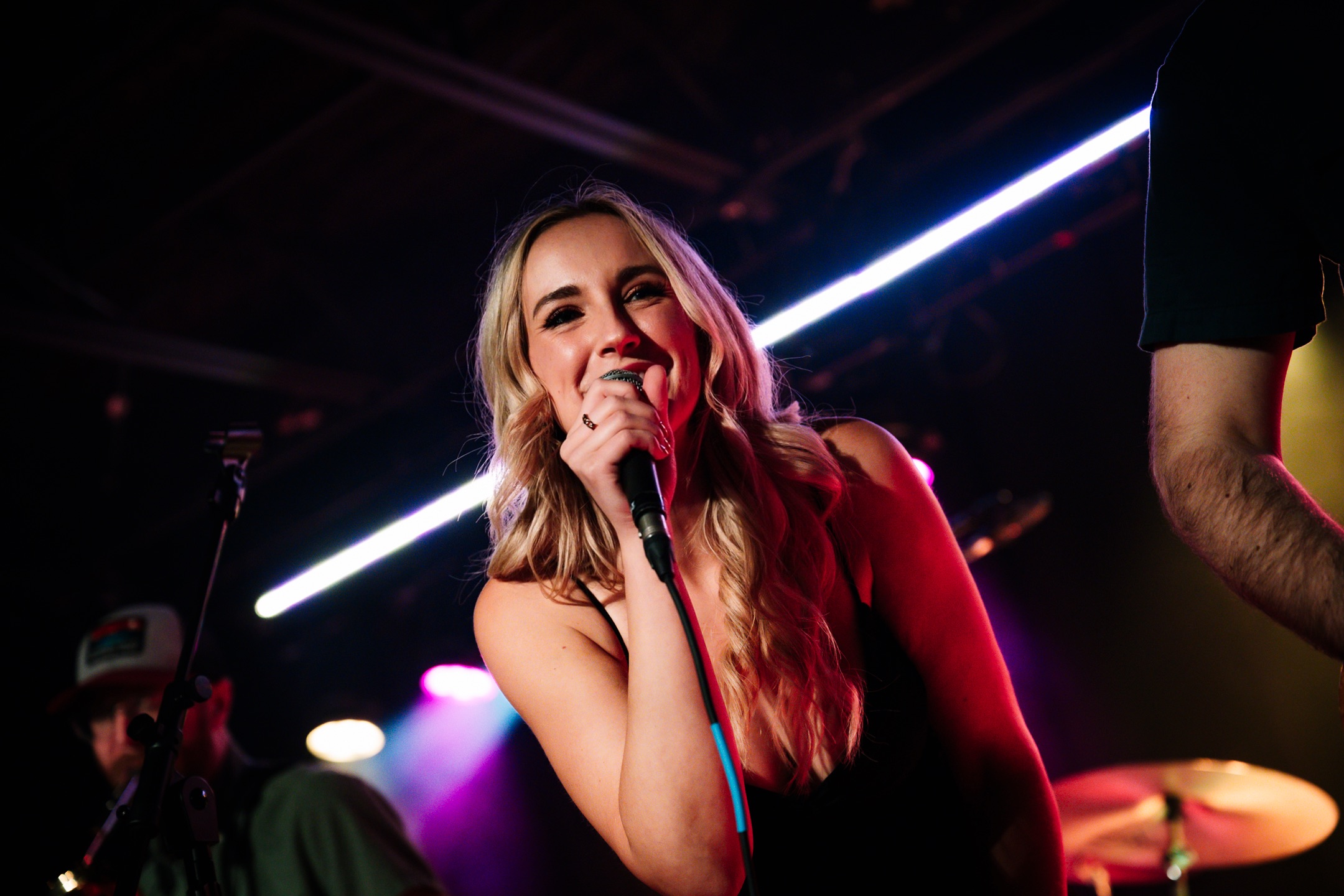 A woman with long blonde hair singing into a microphone on stage, illuminated by colorful stage lights.