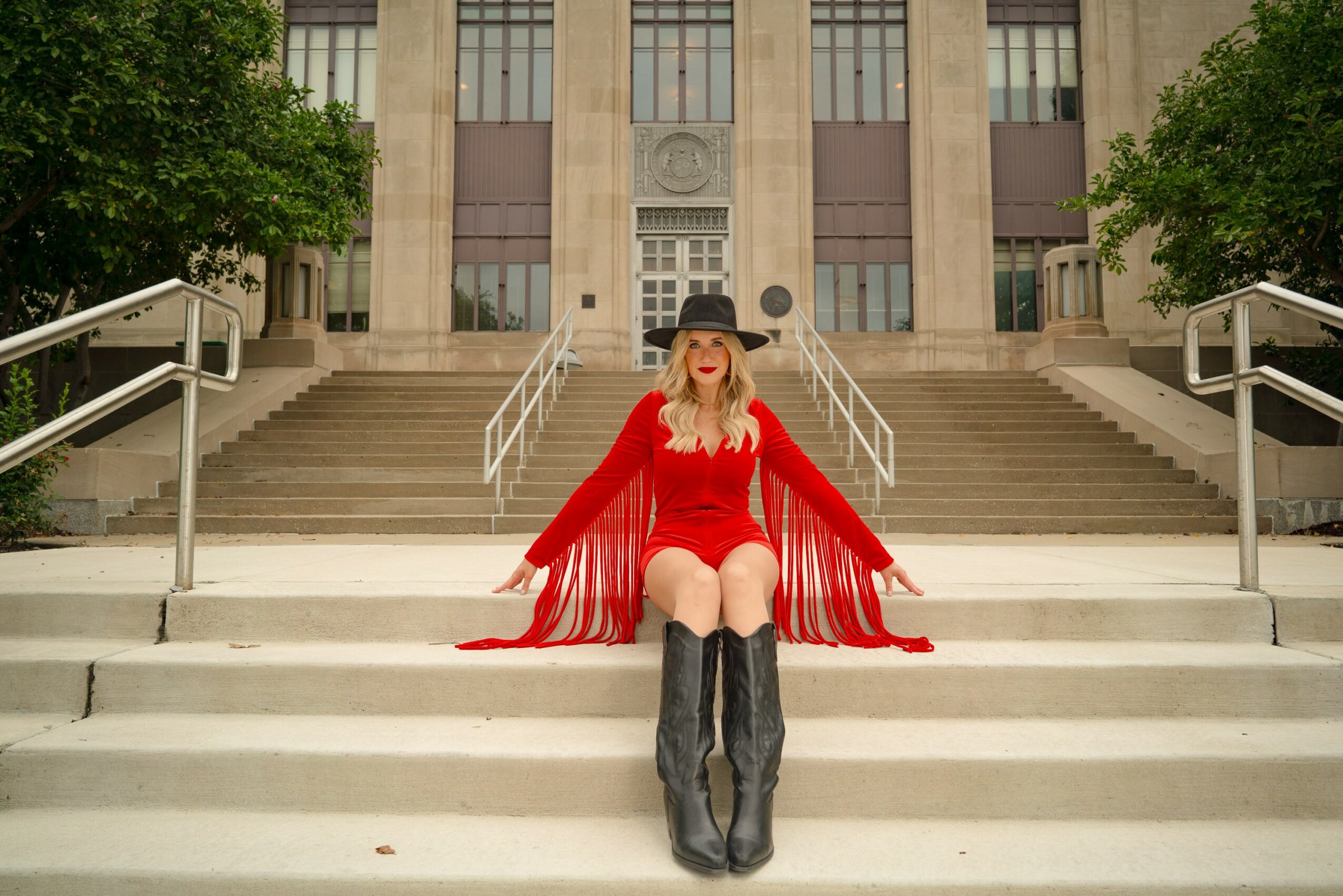 Person sitting on stairs in front of a building, wearing a red dress, black hat, and tall black boots.