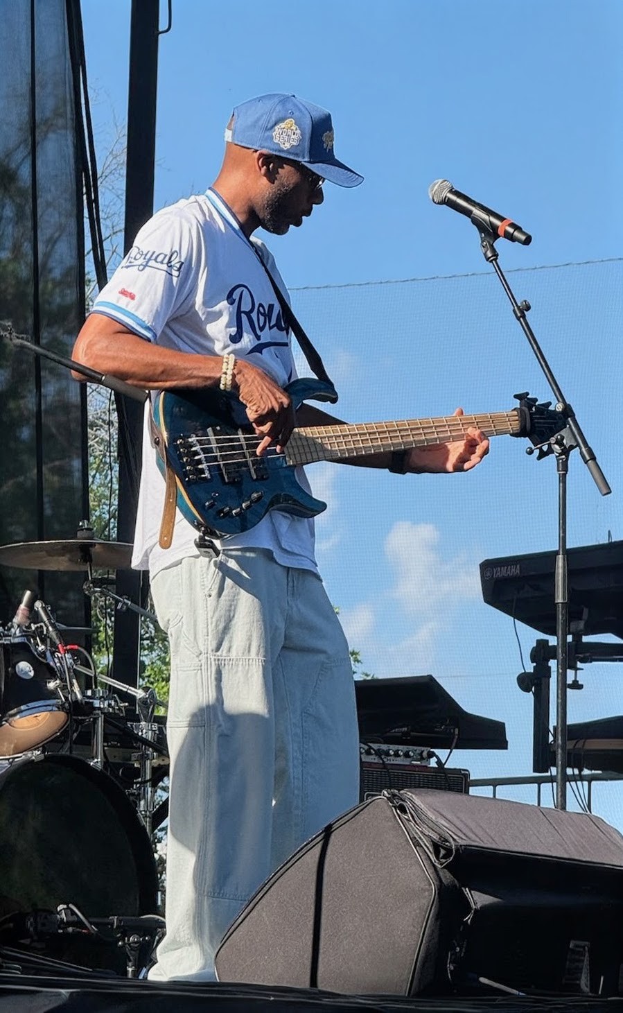 Male musician playing electric guitar on stage outdoors, wearing a blue cap and sunglasses, with a drum set and keyboard nearby.