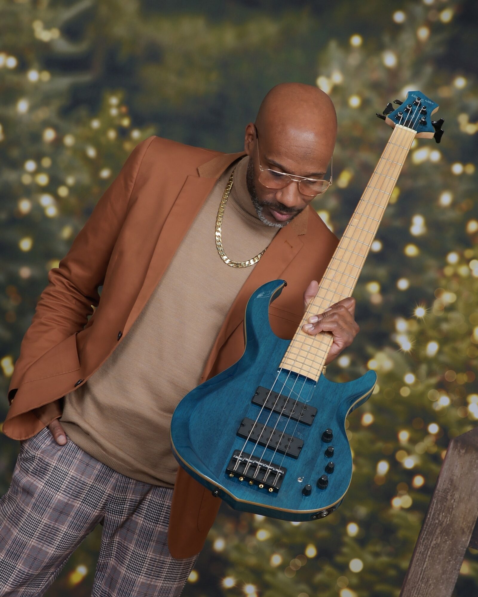 Man holding a blue electric bass guitar, looking down, with a blurred festive background of lights and trees.