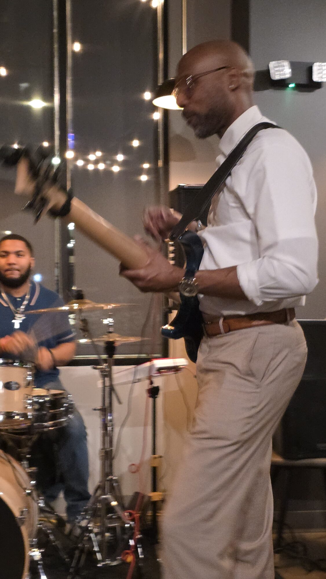 Man playing guitar in a room with drums and another person in the background, illuminated by string lights outside.