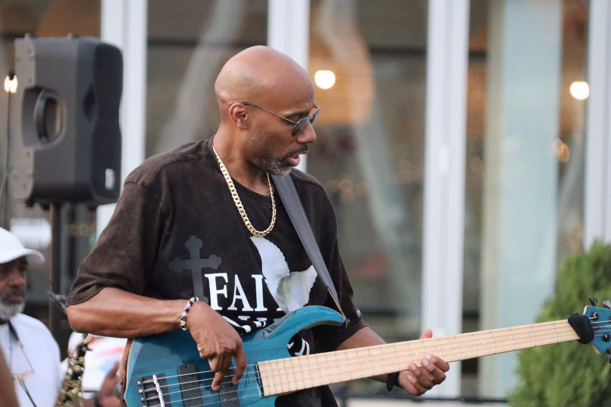 Man playing a blue electric guitar outdoors, wearing glasses, a black T-shirt, and a gold chain, with a speaker nearby.