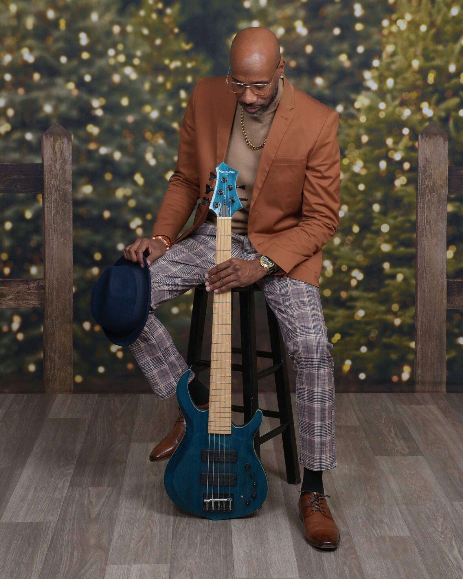 Man sitting on a stool holding a blue bass guitar, with a festive background of decorated trees and lights.
