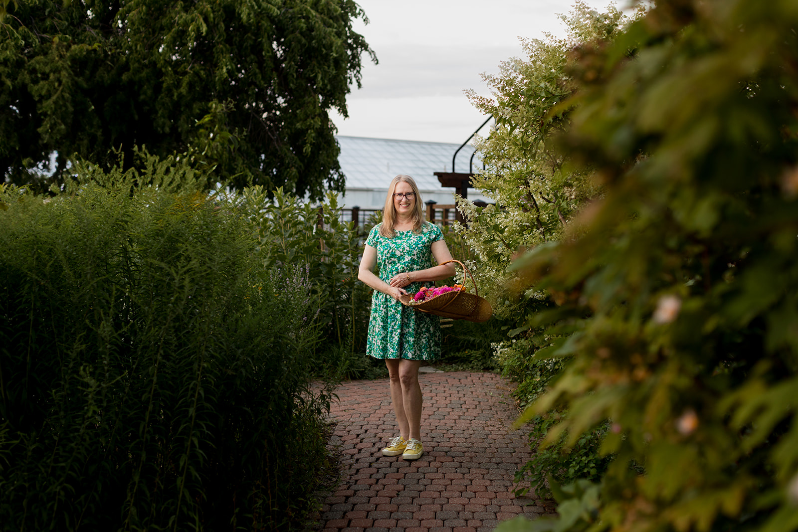 Girl in a green dress holding a basket on a garden path, surrounded by greenery and trees.