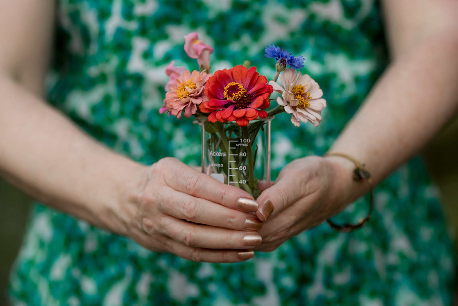 Person holding a glass jar with colorful flowers, wearing a green patterned dress, hands visible, background blurred.