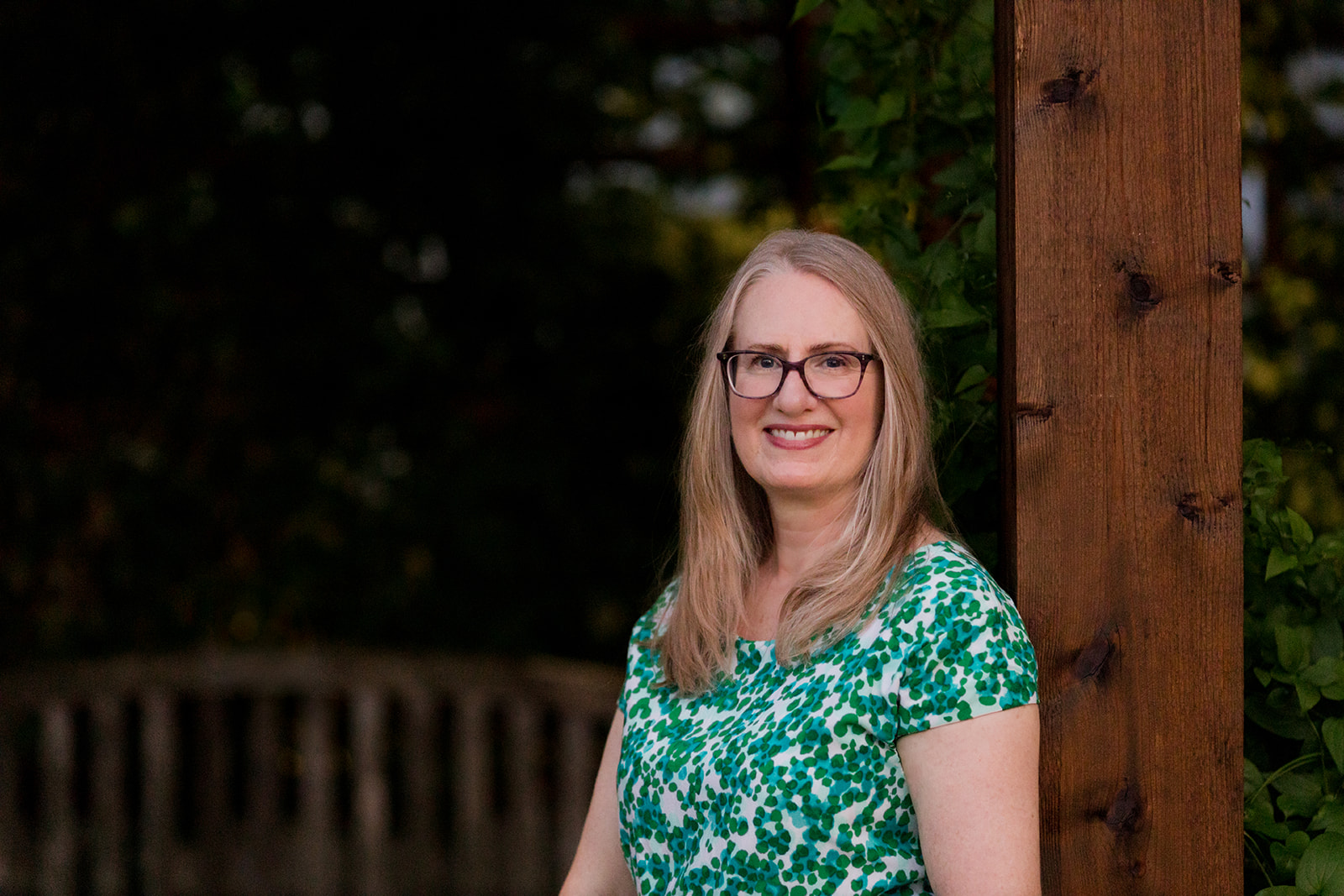 Woman with glasses and long blonde hair smiling outdoors, standing next to a wooden post, wearing a green patterned top.
