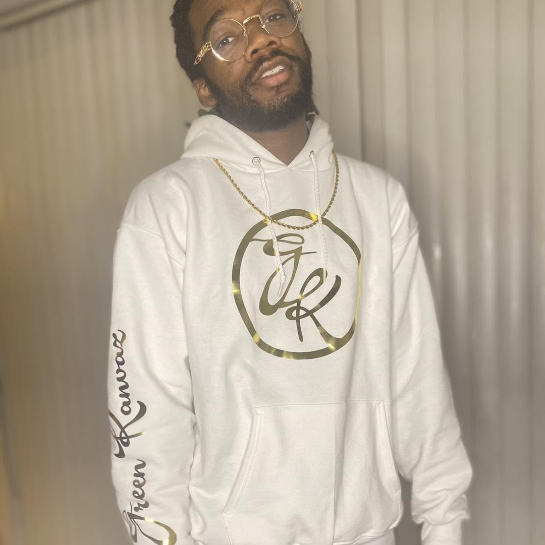 Man with glasses and beard wearing a white hoodie with gold logo, standing in front of vertical blinds.