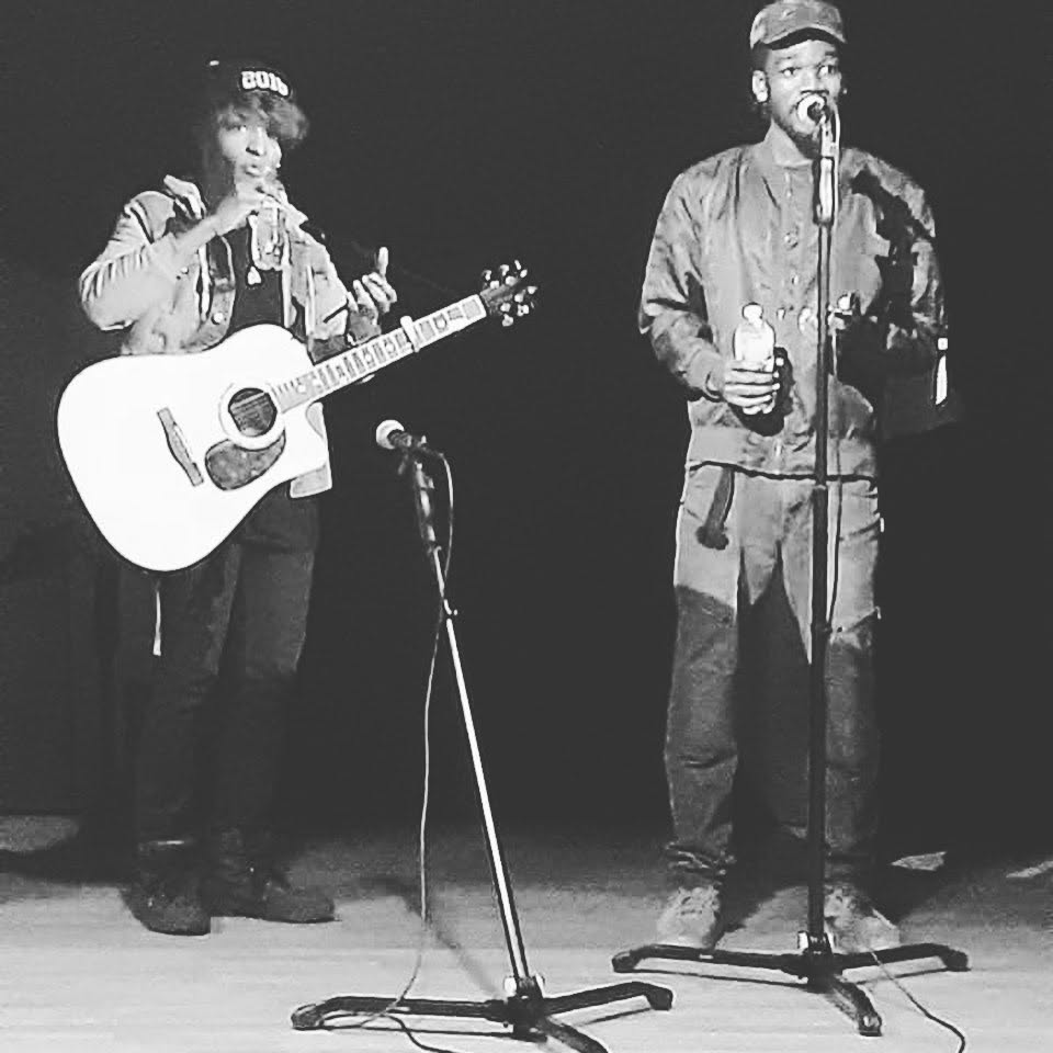 Two people perform on stage, one playing guitar and singing, the other holding a water bottle and speaking into a microphone.