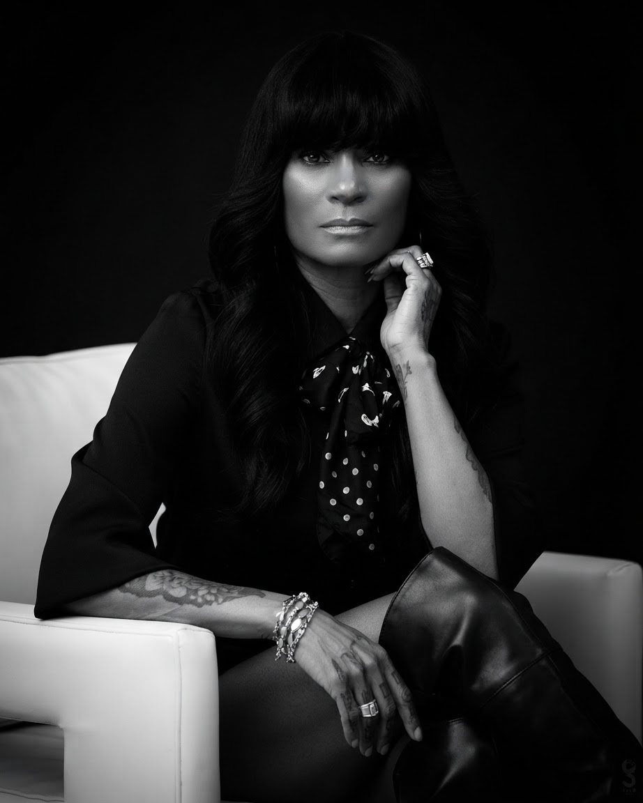 Black and white portrait of a woman with long dark hair, sitting on a white chair, wearing a dark outfit, with her hand near her face.