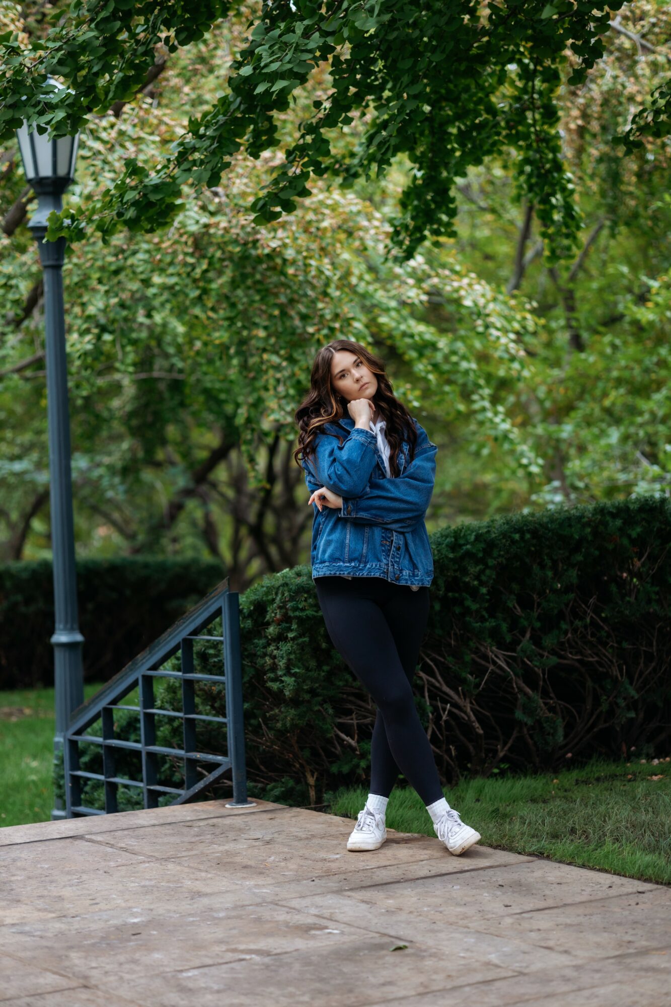 Young woman with long hair in a blue jacket and black pants stands on a sidewalk near a lamppost and greenery.