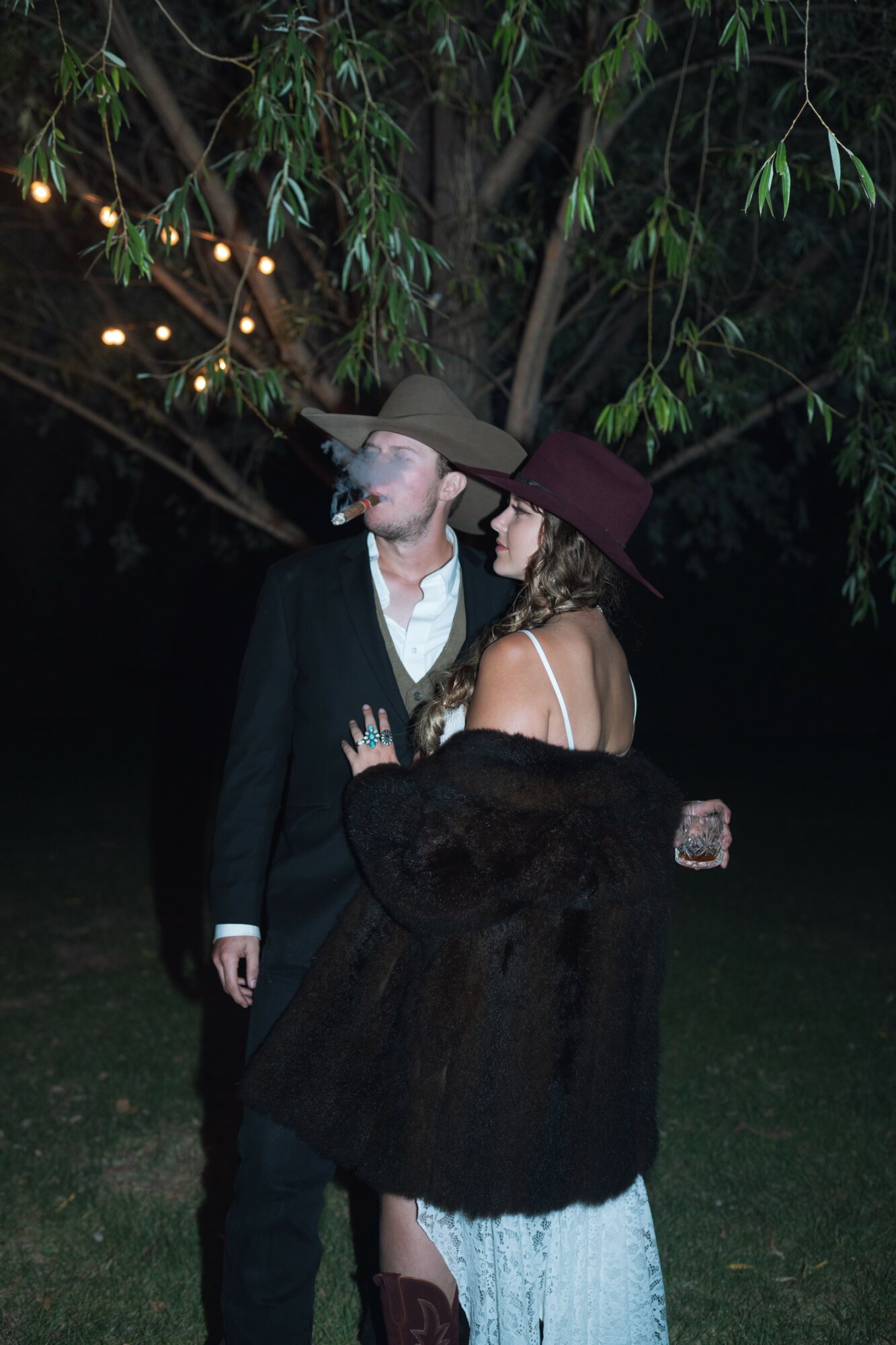 A man and woman in vintage clothing stand close together outdoors at night, under a tree with string lights.