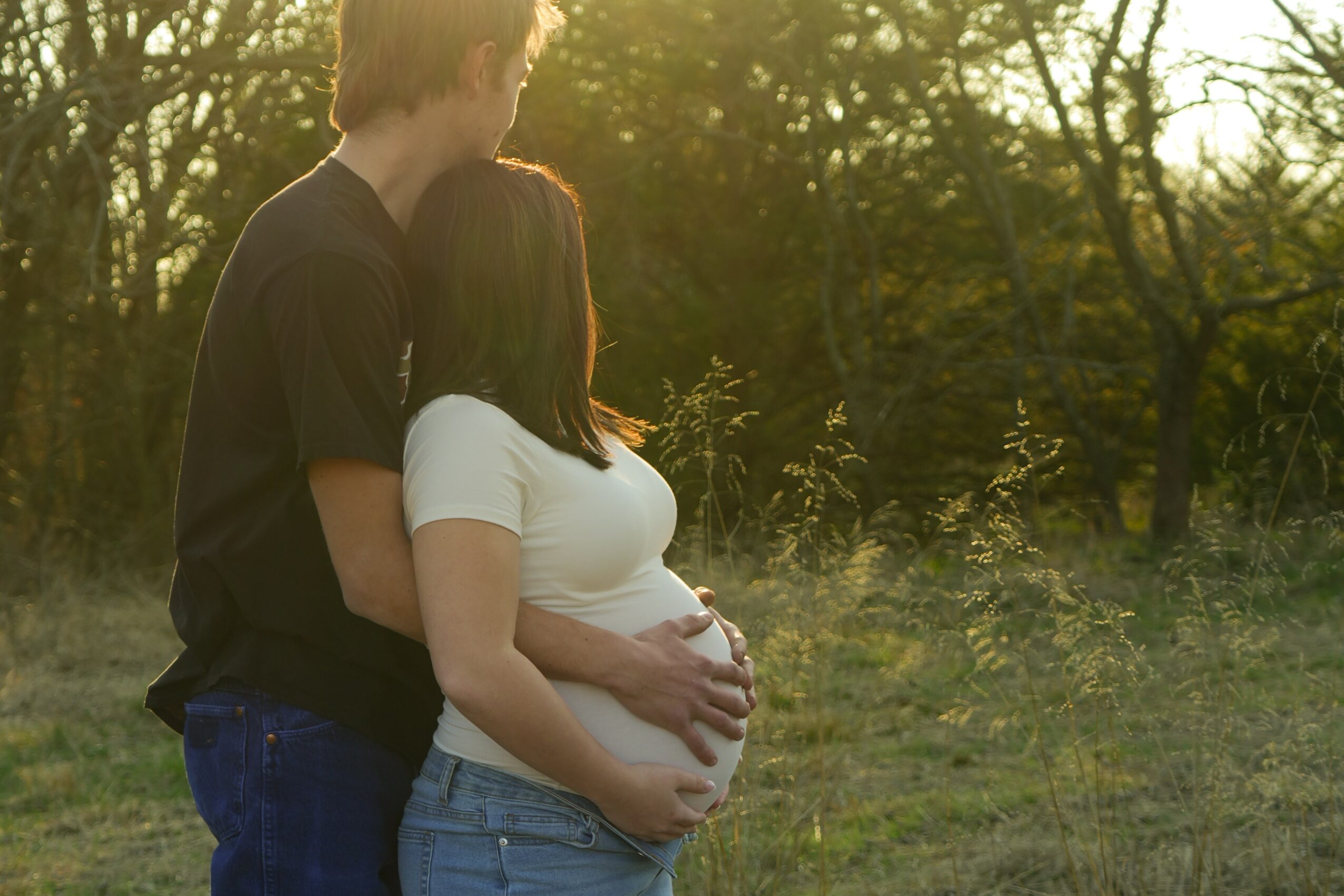 A pregnant woman and a man stand outdoors in a grassy area with trees, embracing at sunset.
