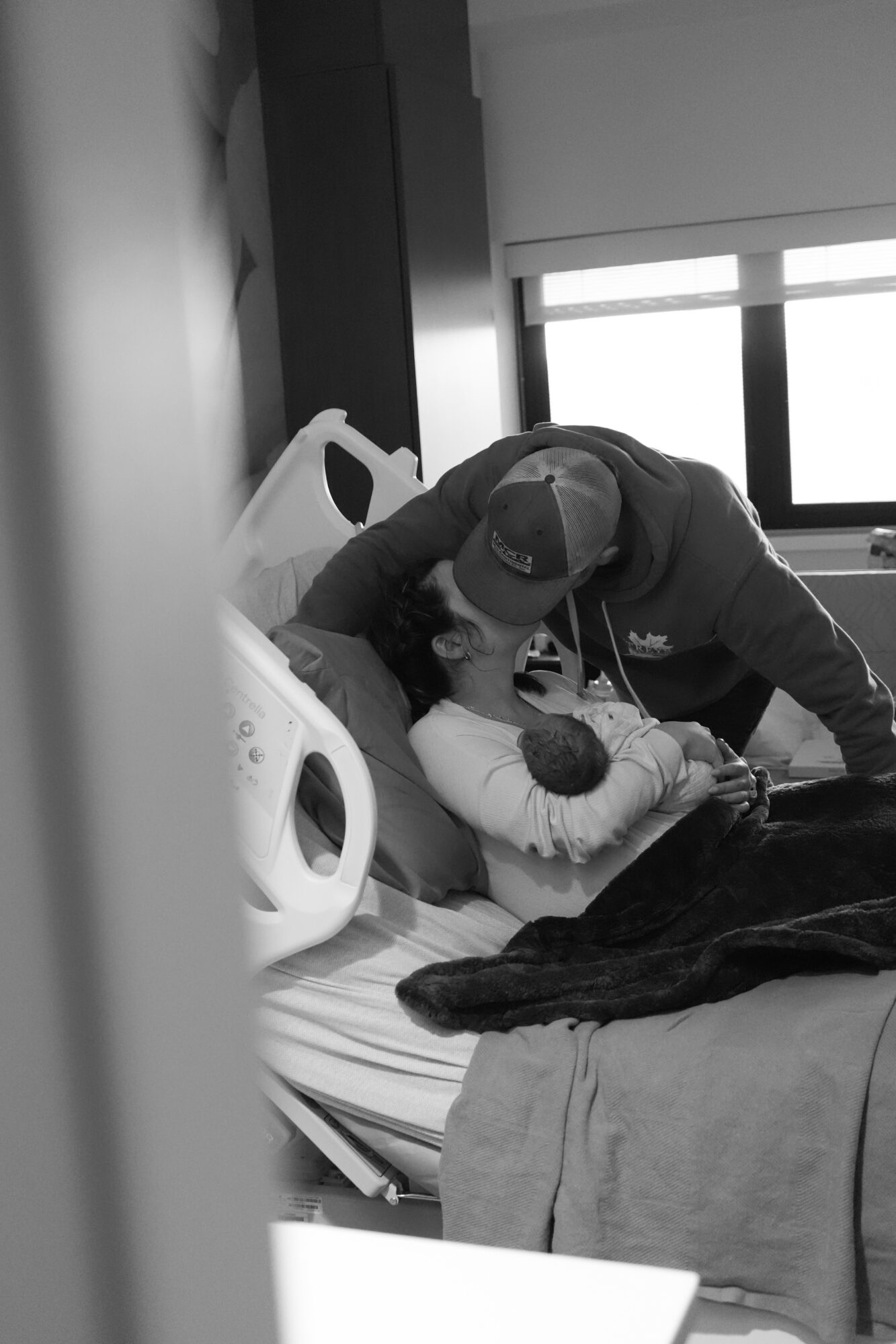Person in hospital bed holding a newborn baby, with a healthcare worker leaning over, in a hospital room.