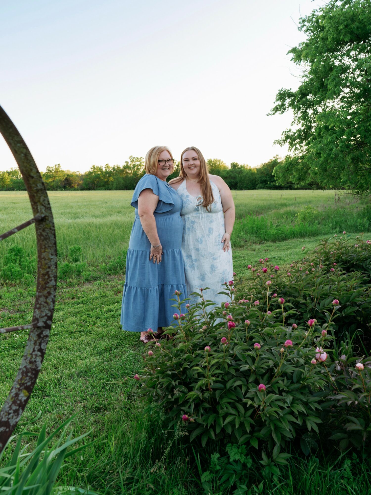 Two women stand outdoors in a grassy field with trees and pink flowers, smiling at the camera.