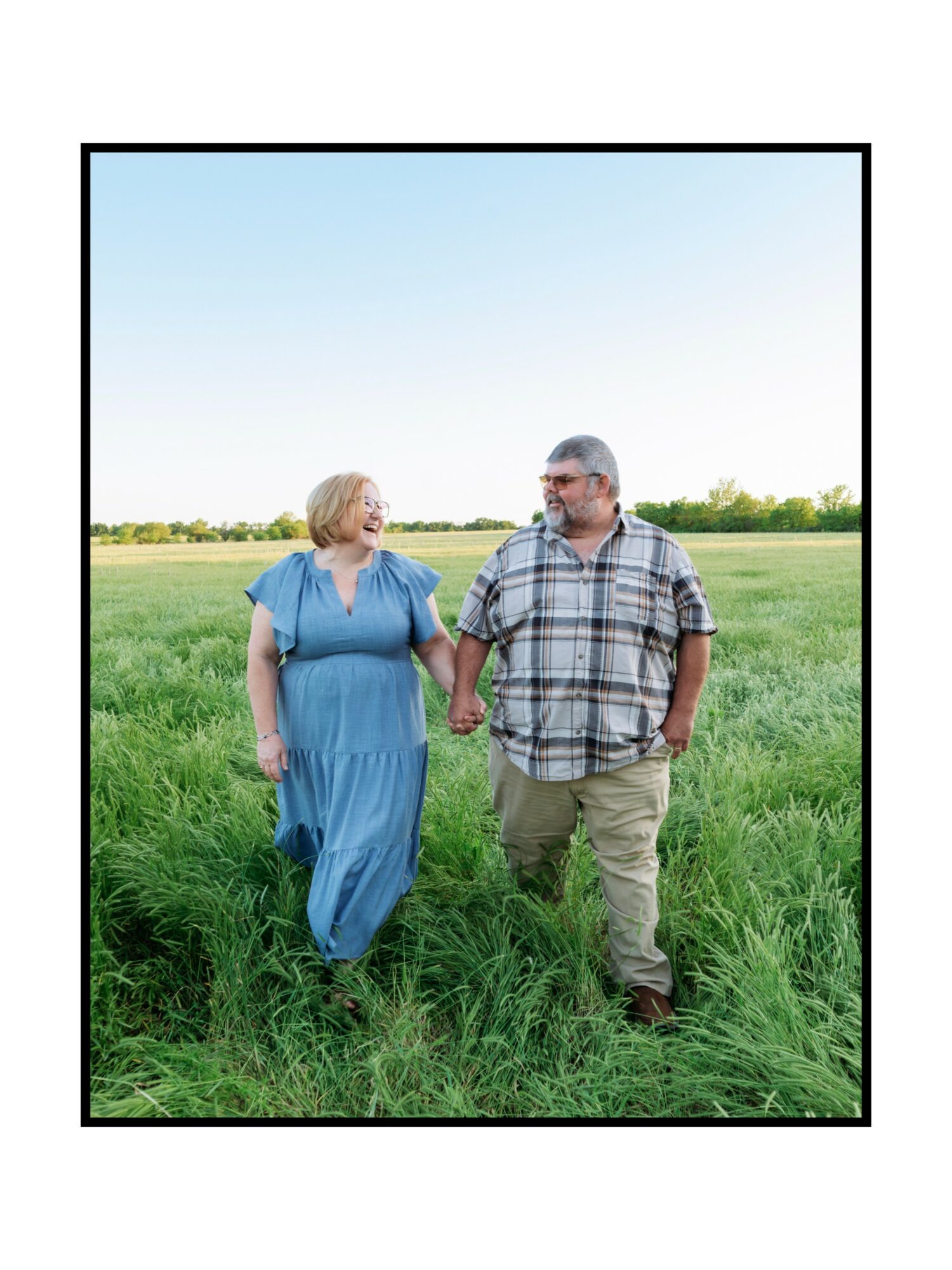 A man and woman walk hand in hand through a grassy field on a clear day.