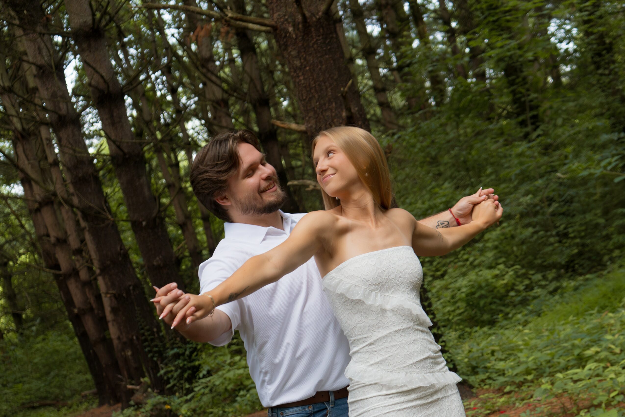 A couple dancing outdoors in a forest, smiling and holding hands, surrounded by green trees and foliage.