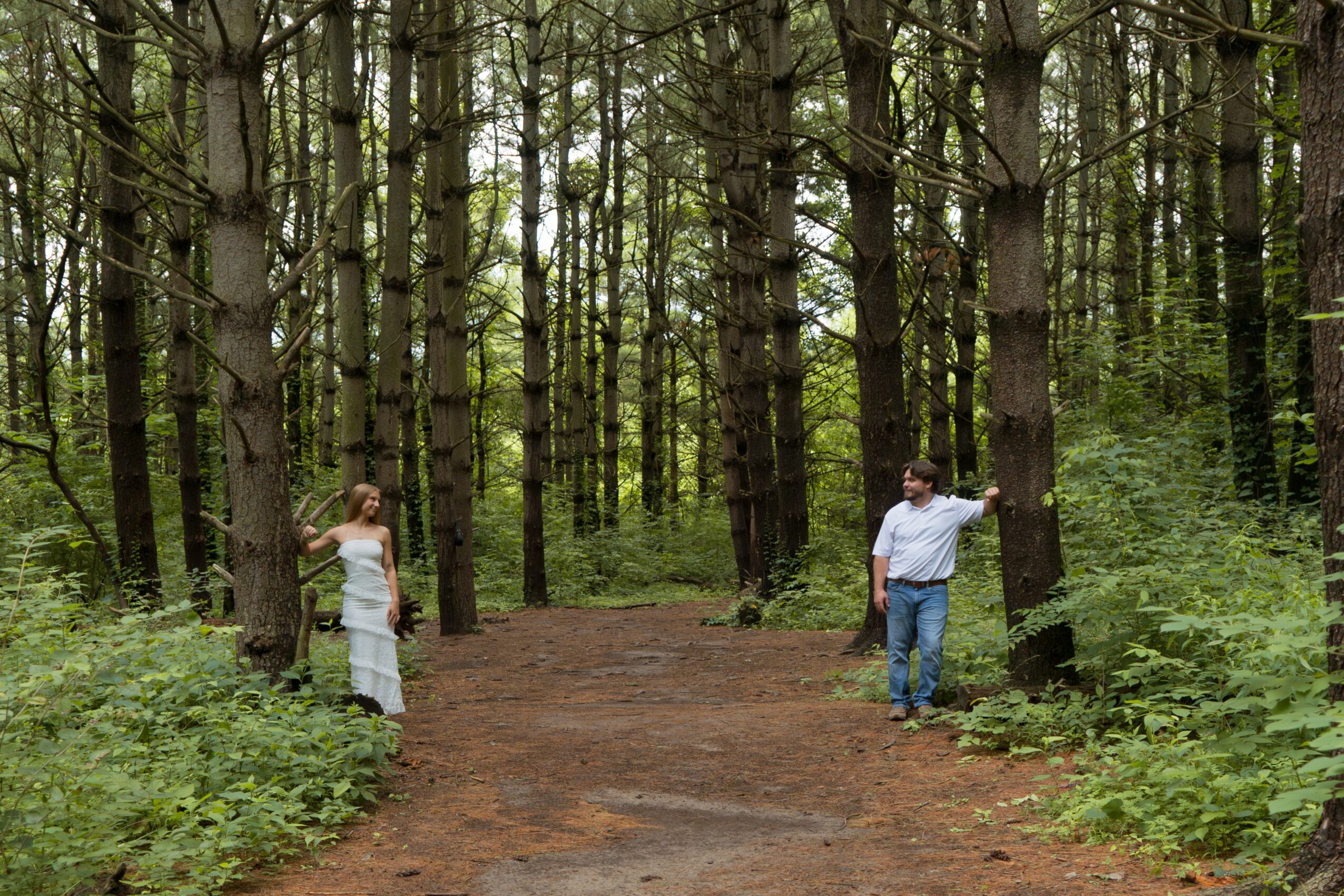 Two people standing in a forest with tall trees and green foliage, facing each other on a dirt path.