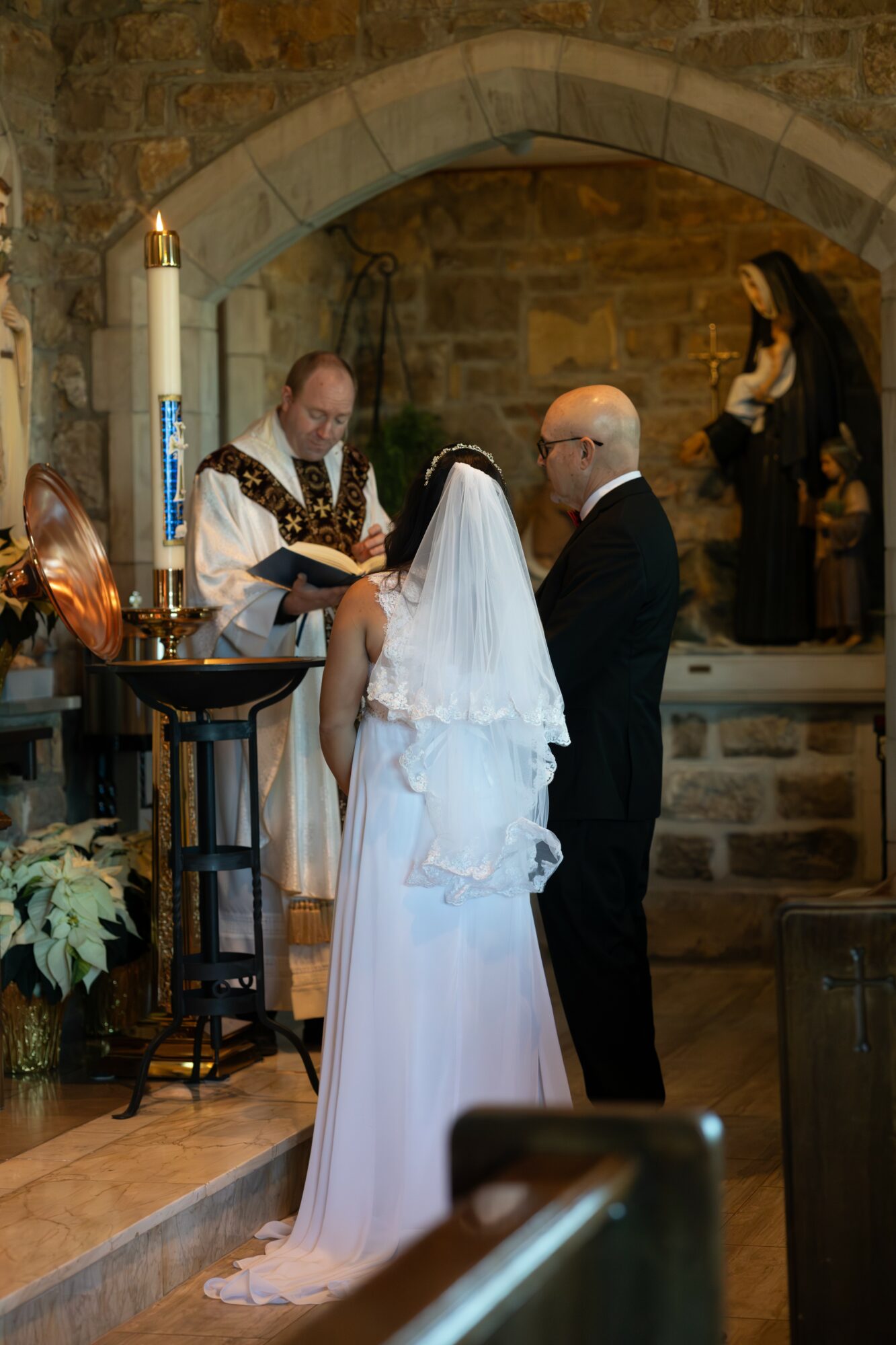 Bride and groom standing before officiant during wedding ceremony in a stone church, with a nun in background.