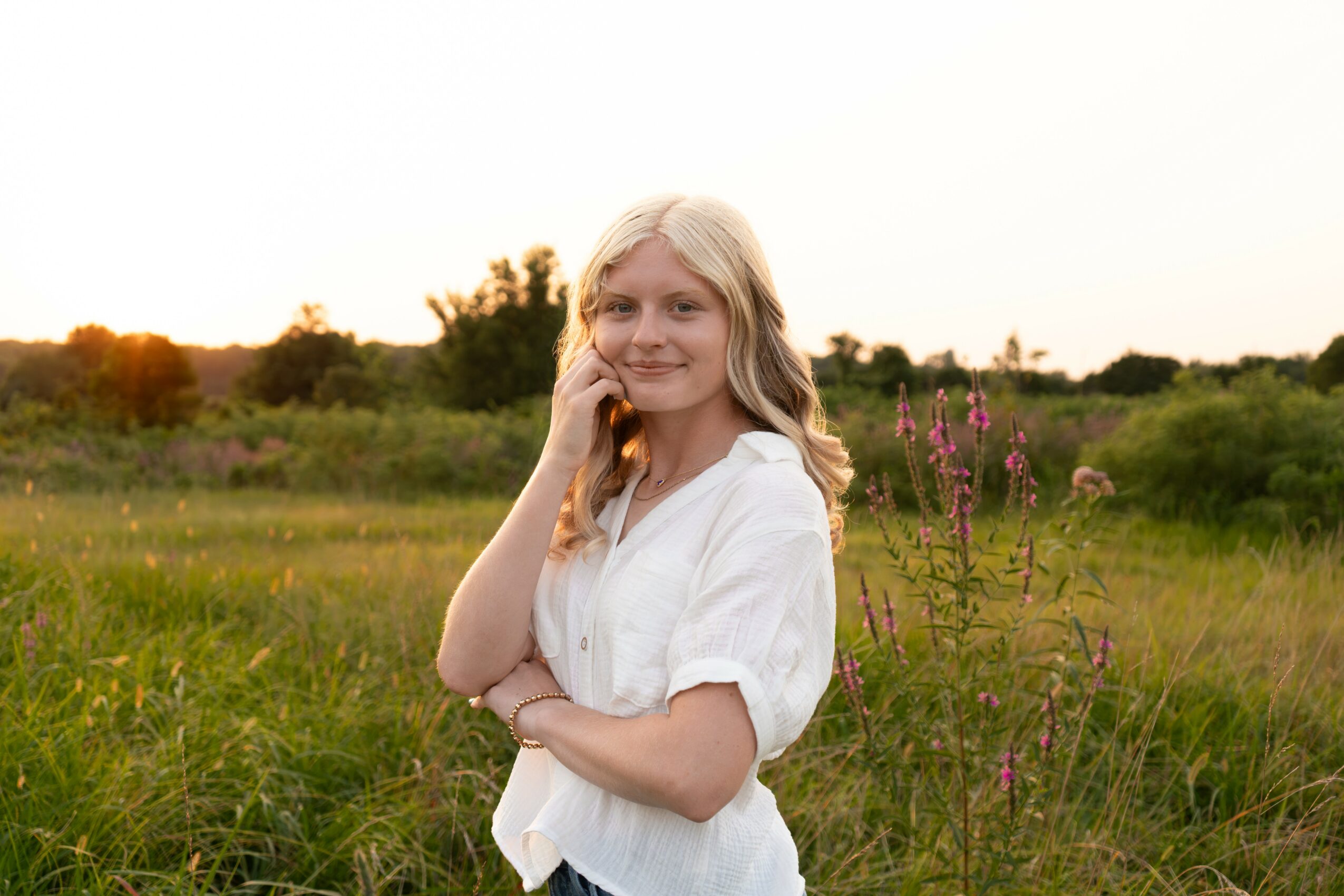 Young woman with blonde hair standing in a grassy field during sunset, smiling and looking at the camera.