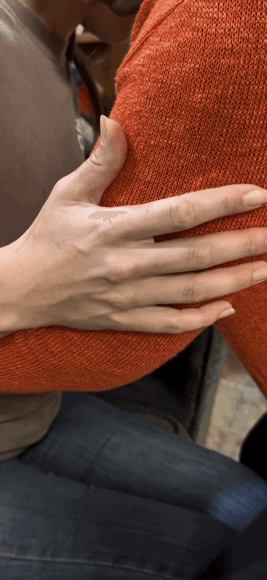 Person's hand resting on another person's arm, both wearing orange clothing, with a background of jeans and a blurred setting.