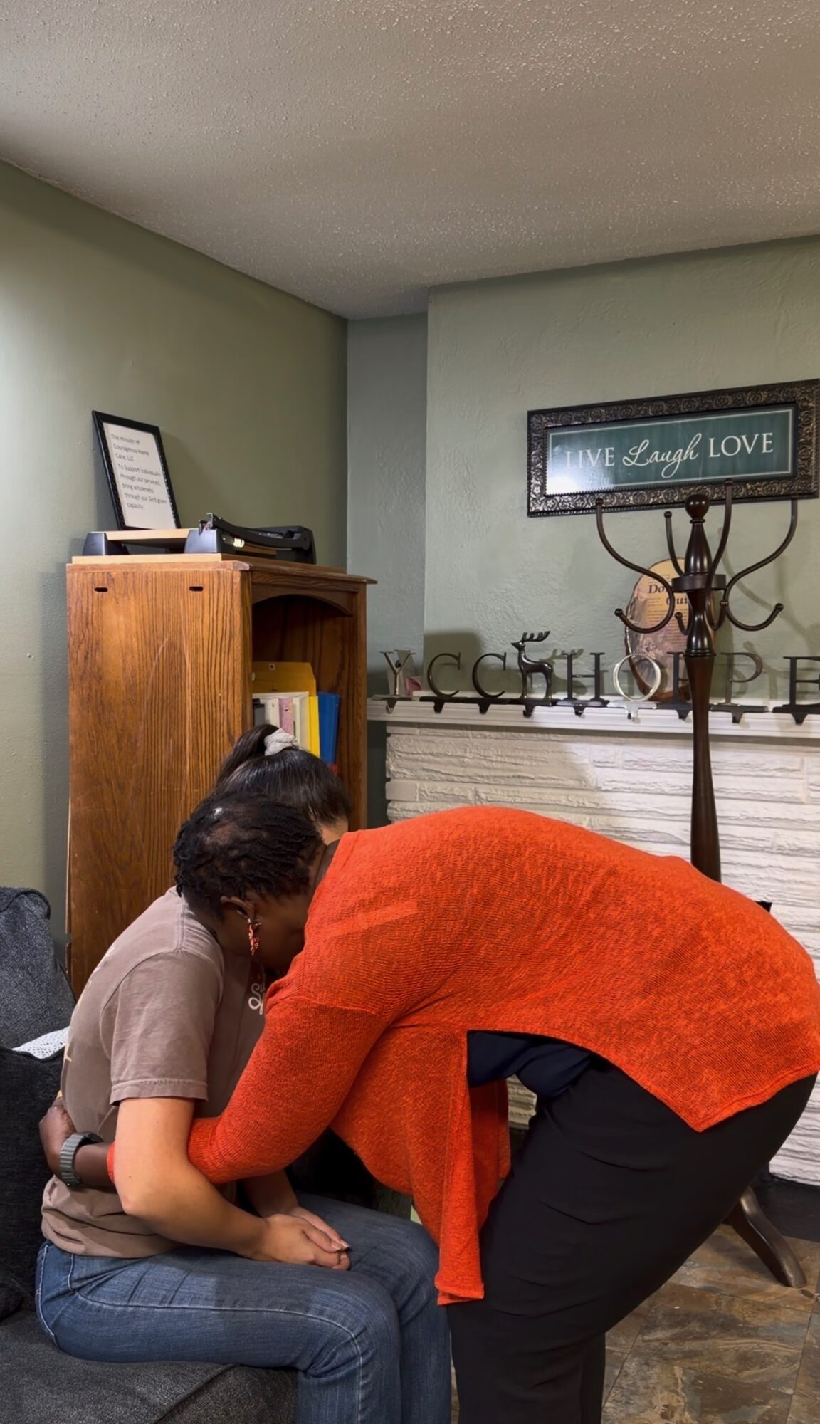 Two people sitting closely, one leaning over the other, in a room with a coat rack and framed wall art.