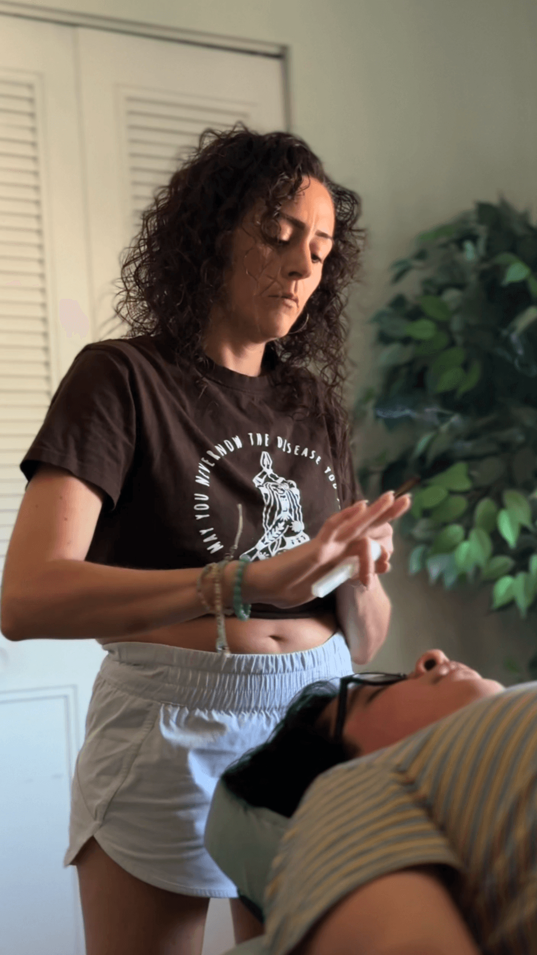 Woman with curly hair looking at her hands, standing near a bed with a person lying down, green plant in background.