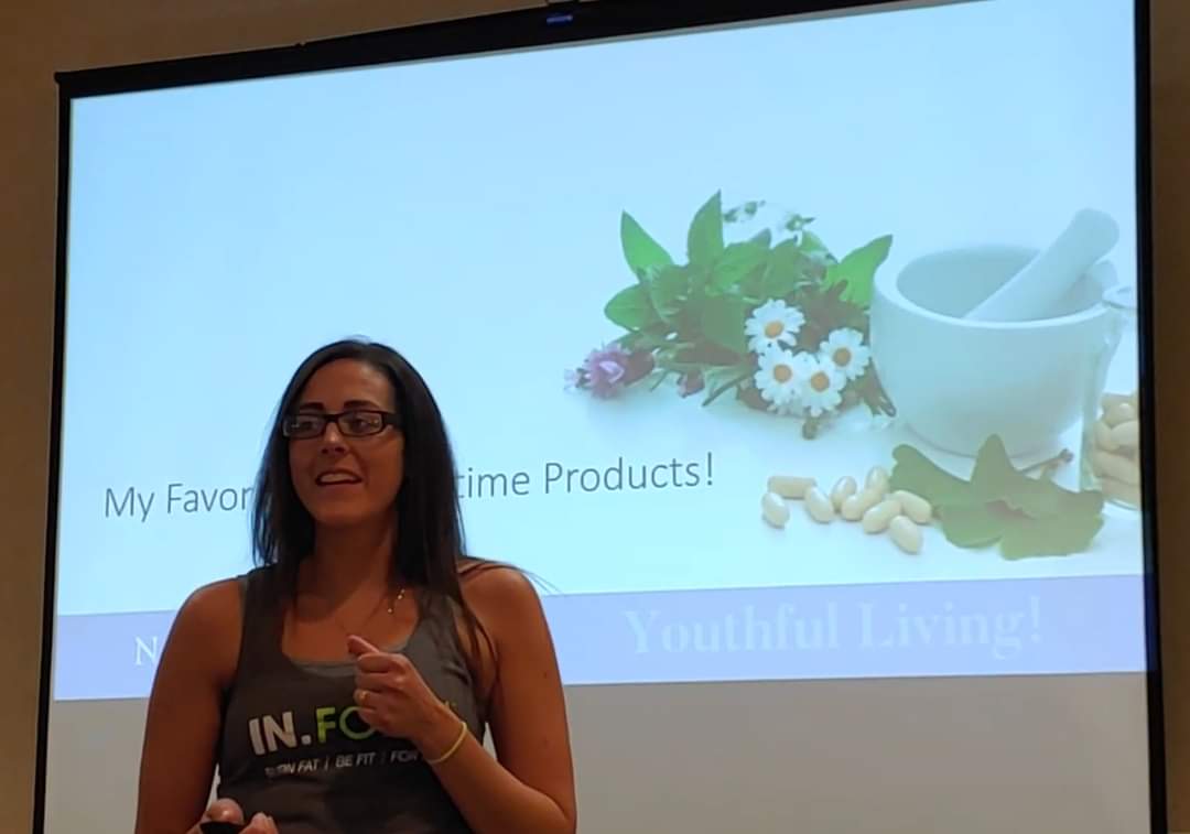 Woman with glasses standing in front of a presentation screen, smiling and gesturing with her hand.