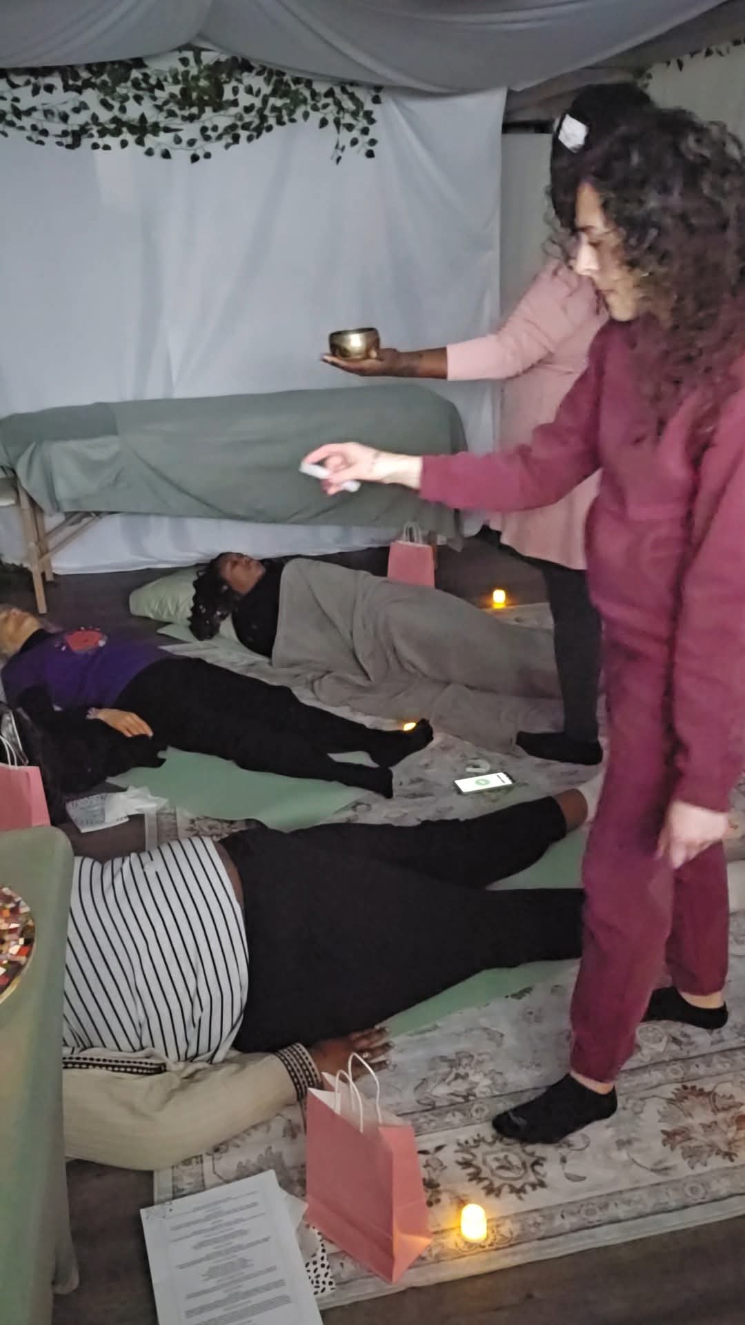 People lying on beds with candles, a woman in a red outfit standing, and another woman holding a bowl, in a decorated room.
