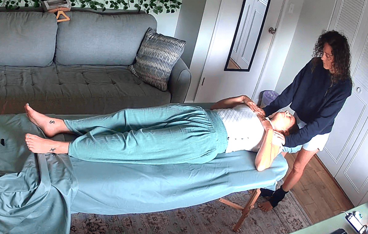 Person lying on a massage table with another person standing beside, holding their arm, in a room with a sofa and door.