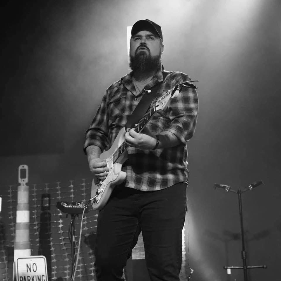 Man with a beard and cap playing electric guitar on stage, with microphones and equipment in background.